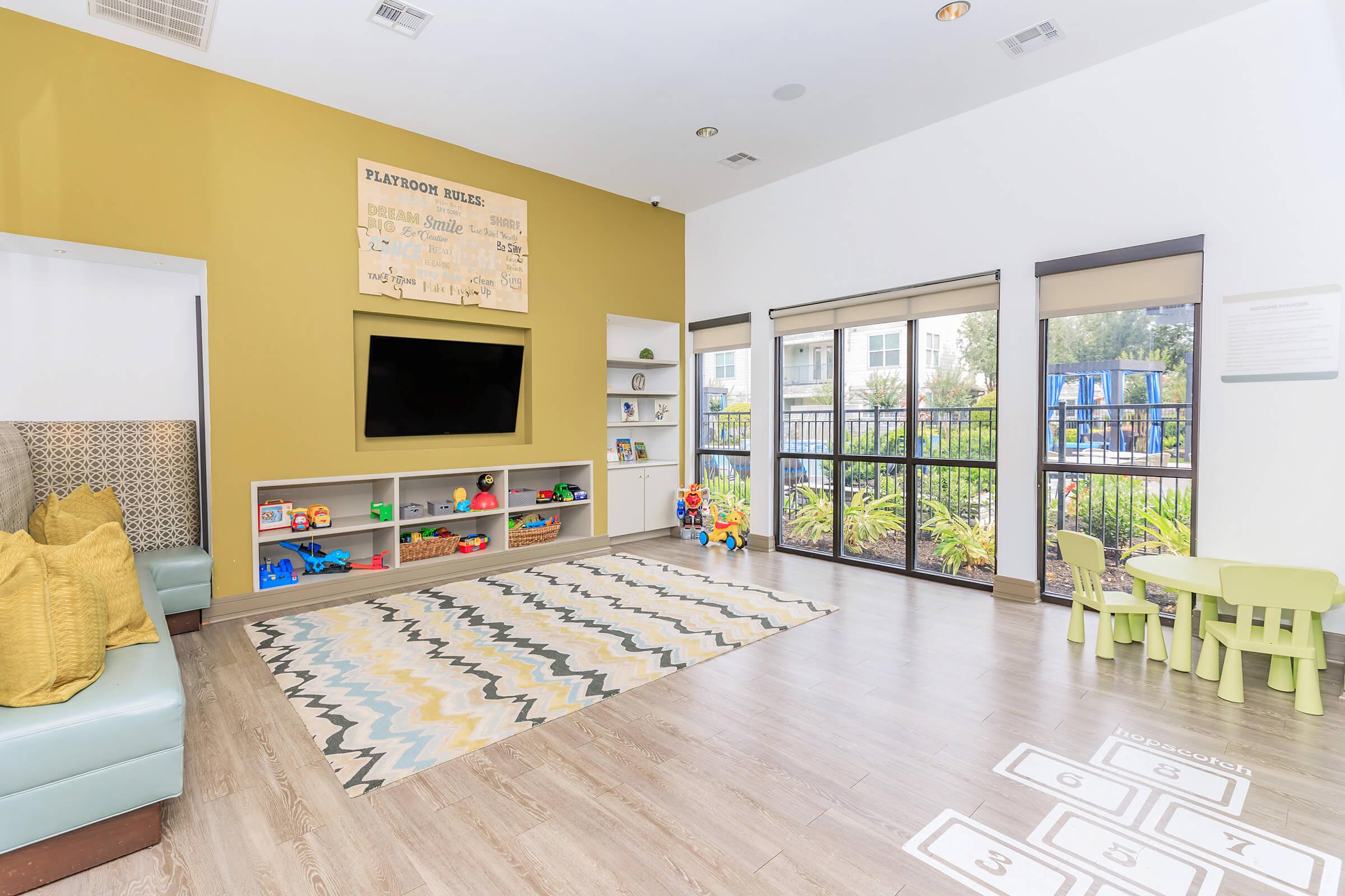 A bright and inviting children's playroom featuring a TV mounted on a yellow wall, a cozy seating area with pillows, colorful toys on shelves, and a patterned rug. There's a small green table with chairs for kids and a view of outdoor playground equipment visible through the large windows.