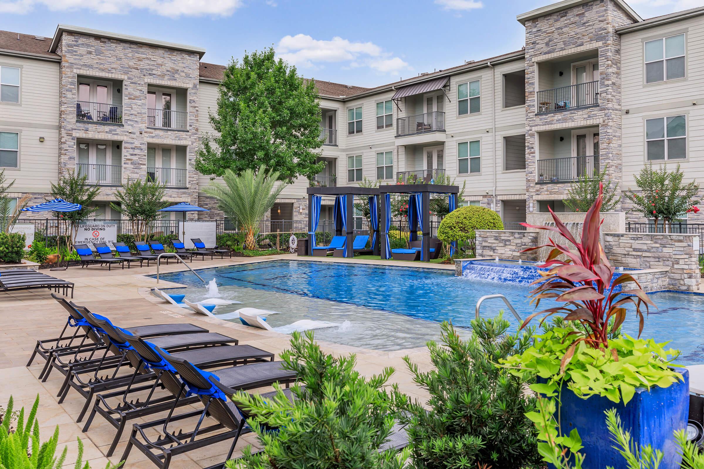 A sunny outdoor pool area at an apartment complex, featuring a large swimming pool with a fountain, surrounded by lounge chairs and lush greenery. In the background, there are modern residential buildings with balconies, and a shaded gazebo near the pool. Blue umbrellas provide shade.