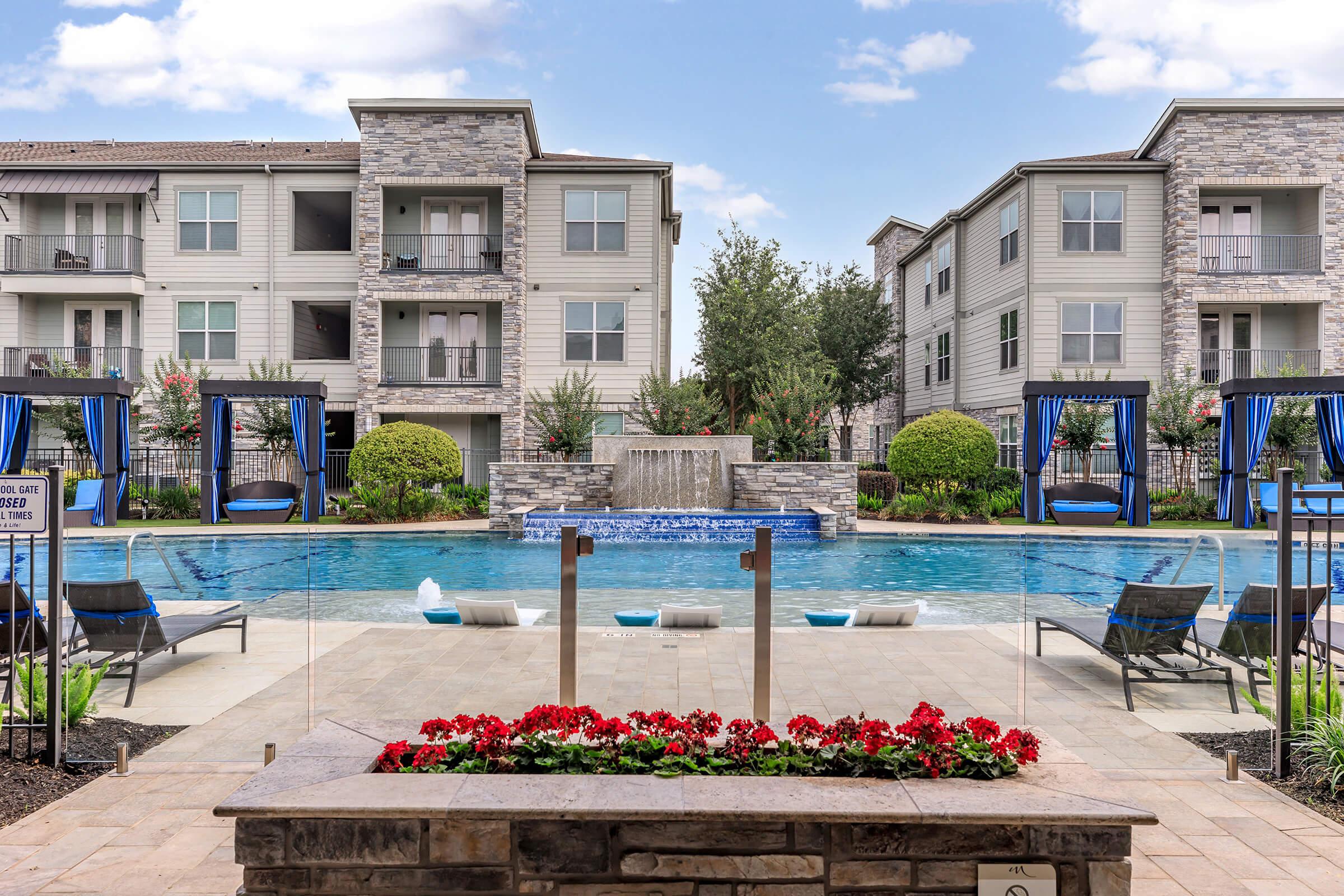 A serene apartment complex pool area featuring a large sparkling blue pool, surrounded by lush landscaping, stone waterfalls, and cozy cabanas. Vibrant flowers in the foreground add color, while modern buildings frame the scene against a partly cloudy sky.