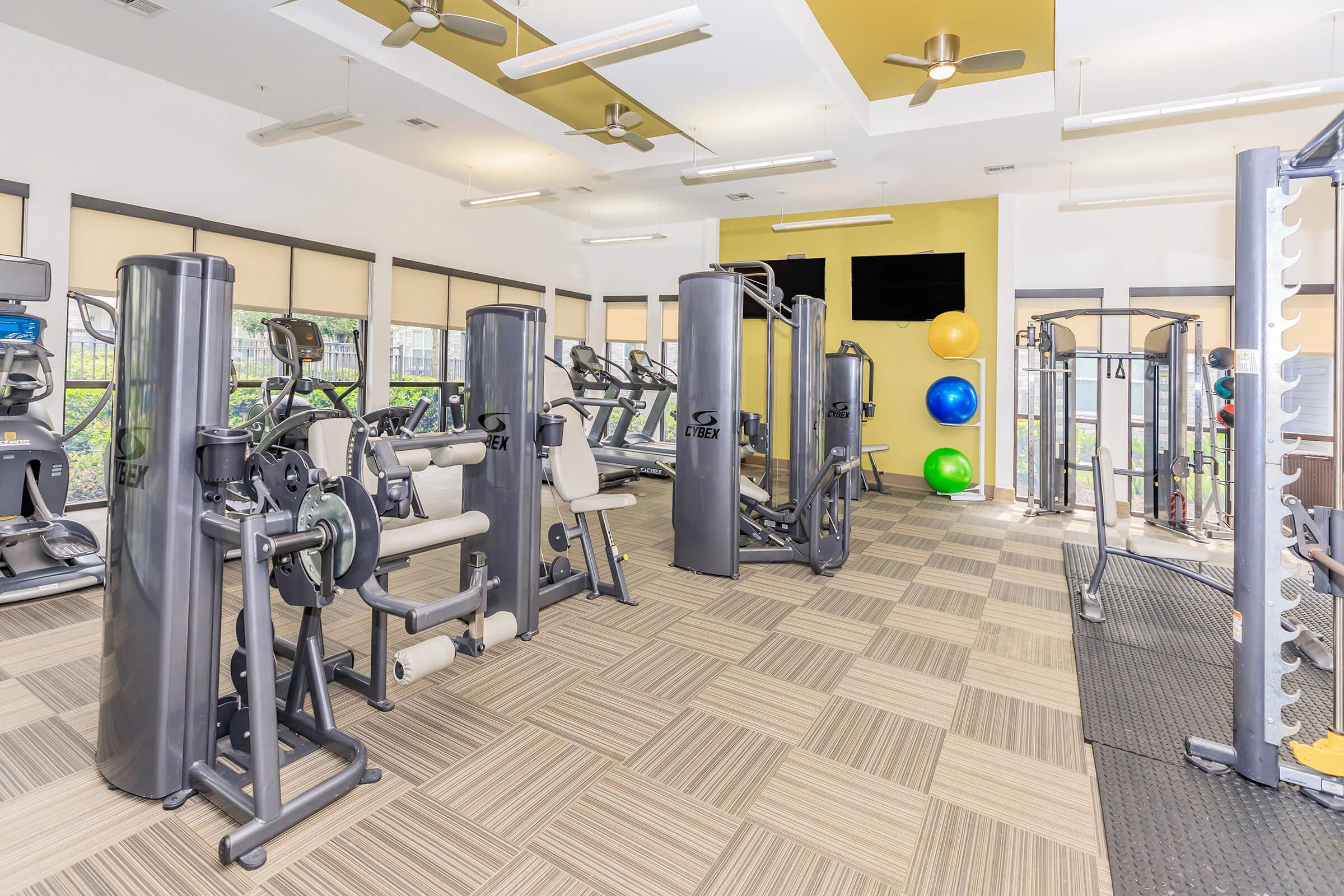 A well-equipped gym interior featuring various exercise machines, cardio equipment, and fitness balls. The space has natural light from large windows, with a contemporary design and tan-colored flooring, creating an inviting atmosphere for workout sessions.