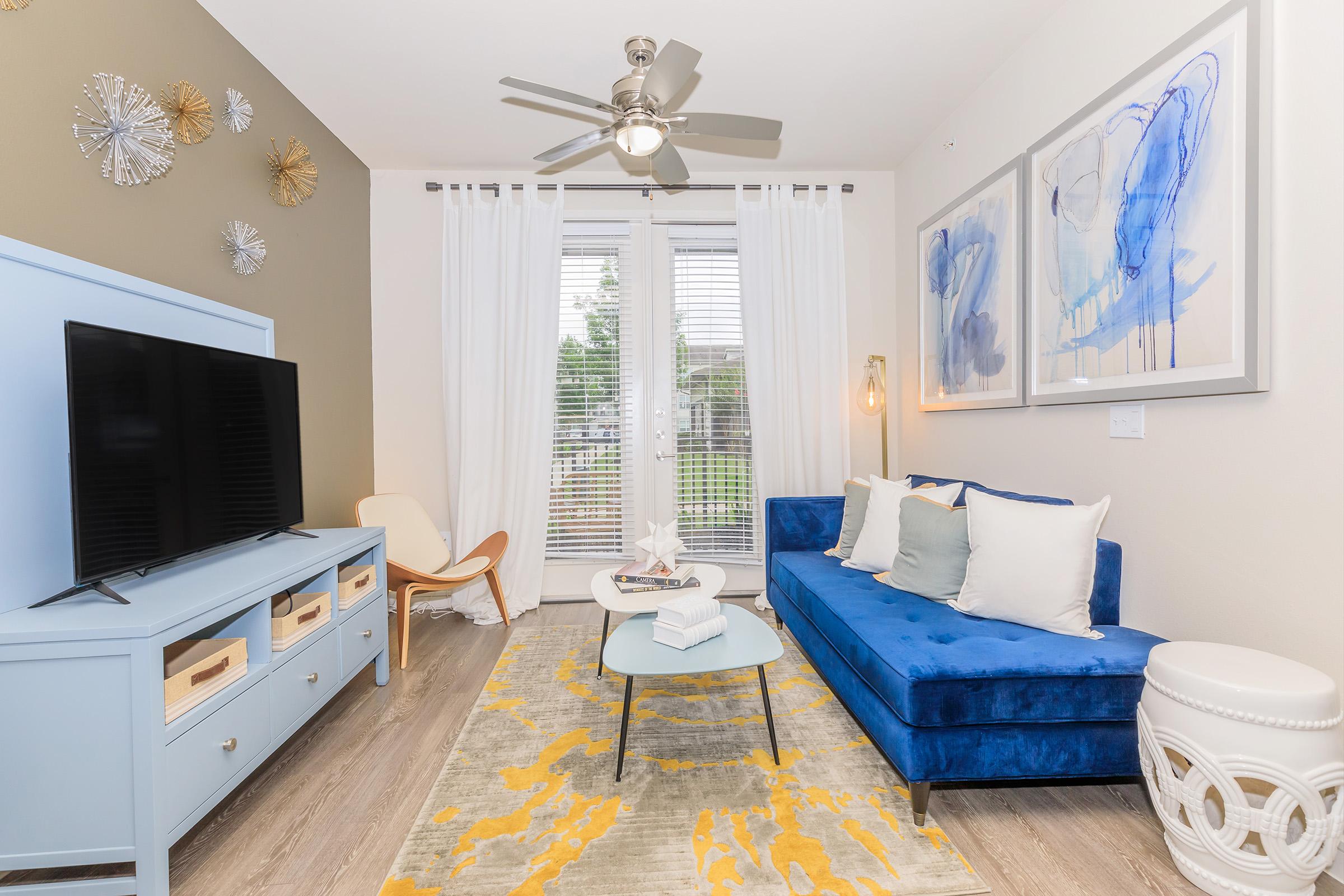 A modern living room featuring a blue velvet couch, a light blue TV stand with storage, and a circular coffee table on a yellow and white patterned rug. Large windows with sheer curtains illuminate the space, complemented by decorative wall art and a ceiling fan.