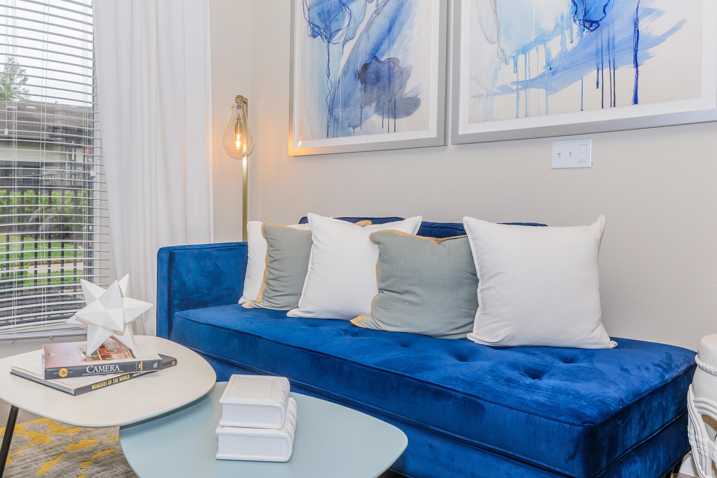 A modern living room featuring a blue velvet sofa with multiple decorative pillows, a side table with a magazine and white decorative items, and abstract artwork on the walls showcasing blue tones. Natural light comes through a nearby window with blinds.