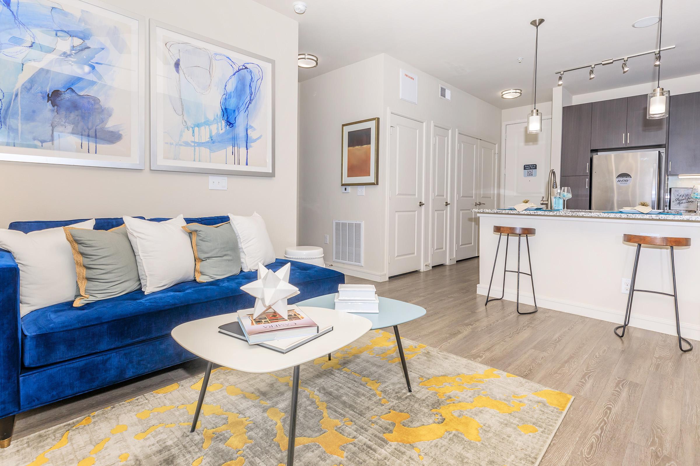 A bright and modern living room featuring a blue couch with white pillows, a light coffee table, and a yellow area rug. There are abstract paintings on the wall, and a kitchen area with stainless steel appliances and bar stools in the background. The space is well-lit with contemporary decor.