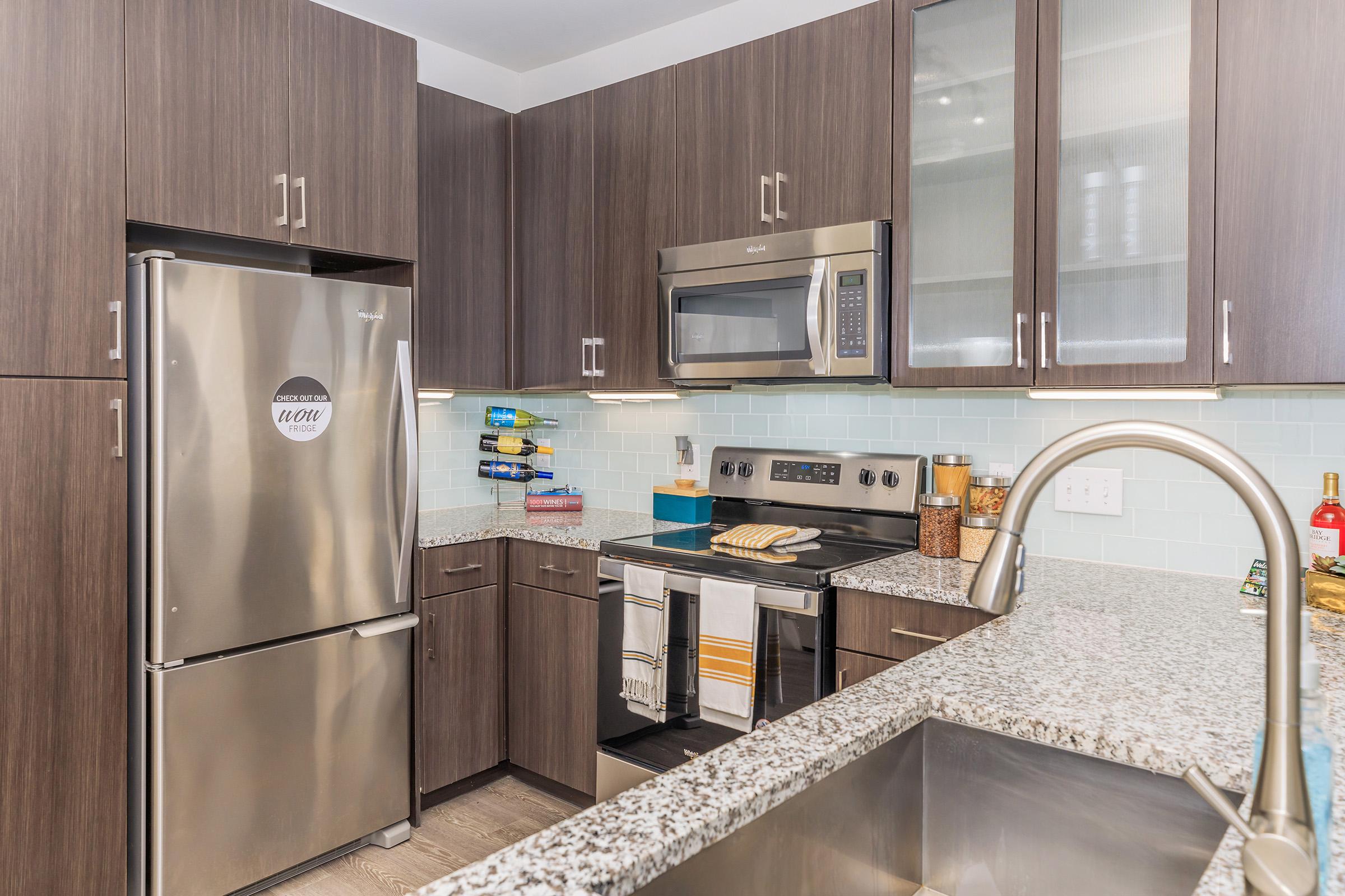 Modern kitchen featuring dark wood cabinetry, a stainless steel refrigerator, and a microwave above the stove. The countertops are made of grey speckled granite, and there are light blue backsplash tiles. A sink with a modern faucet is in the foreground, and various kitchen essentials are neatly organized.