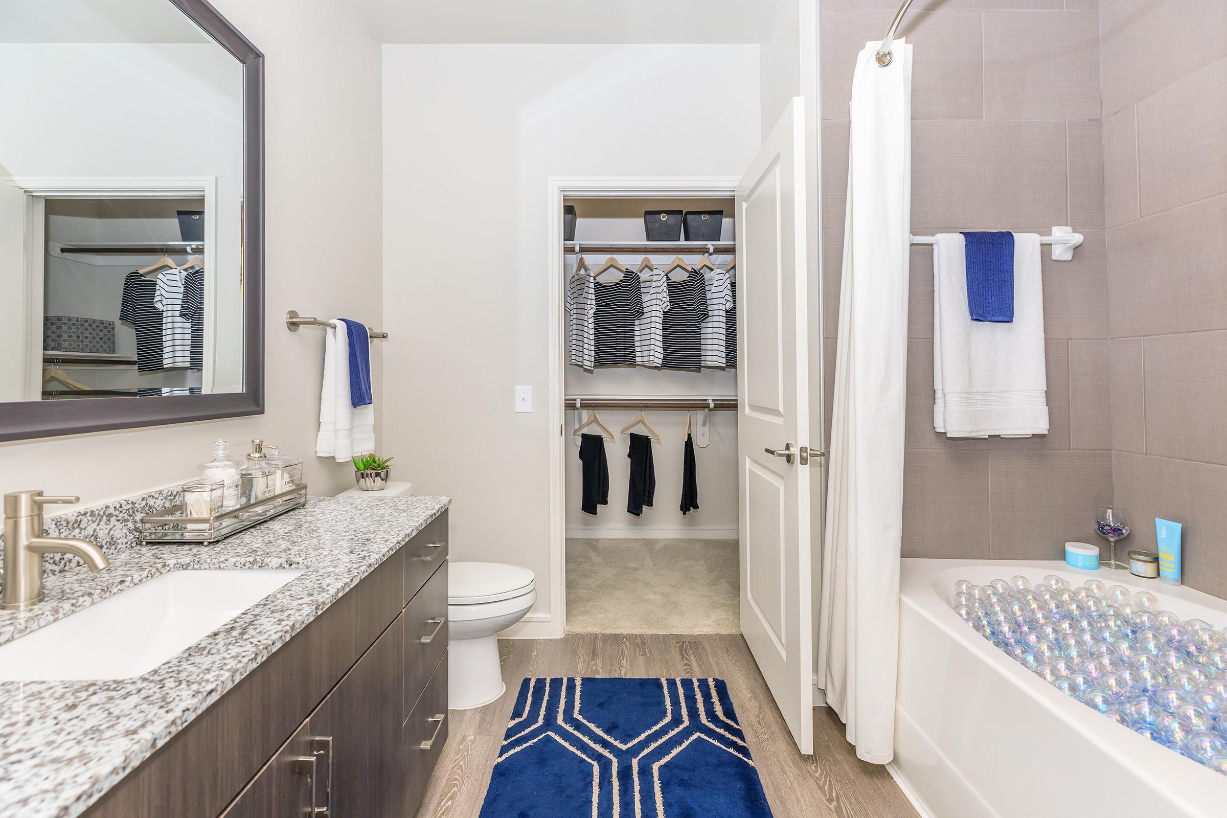 A modern bathroom featuring a white bathtub with colorful bath balls, a sleek vanity with a granite countertop, and a large mirror. The space includes a closet with neatly organized clothes and a blue rug on the floor, enhancing the contemporary aesthetic. Towel bars hold white towels, adding functionality.