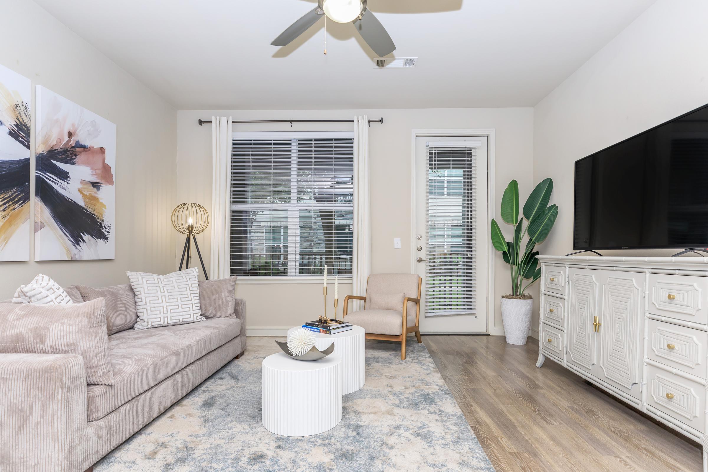A modern living room featuring a cozy beige sofa with decorative pillows, a round white coffee table, and a stylish armchair. There is a large TV mounted on the wall, a window with blinds allowing natural light in, and a plant in a pot. The room has a light color scheme and contemporary artwork on the walls.