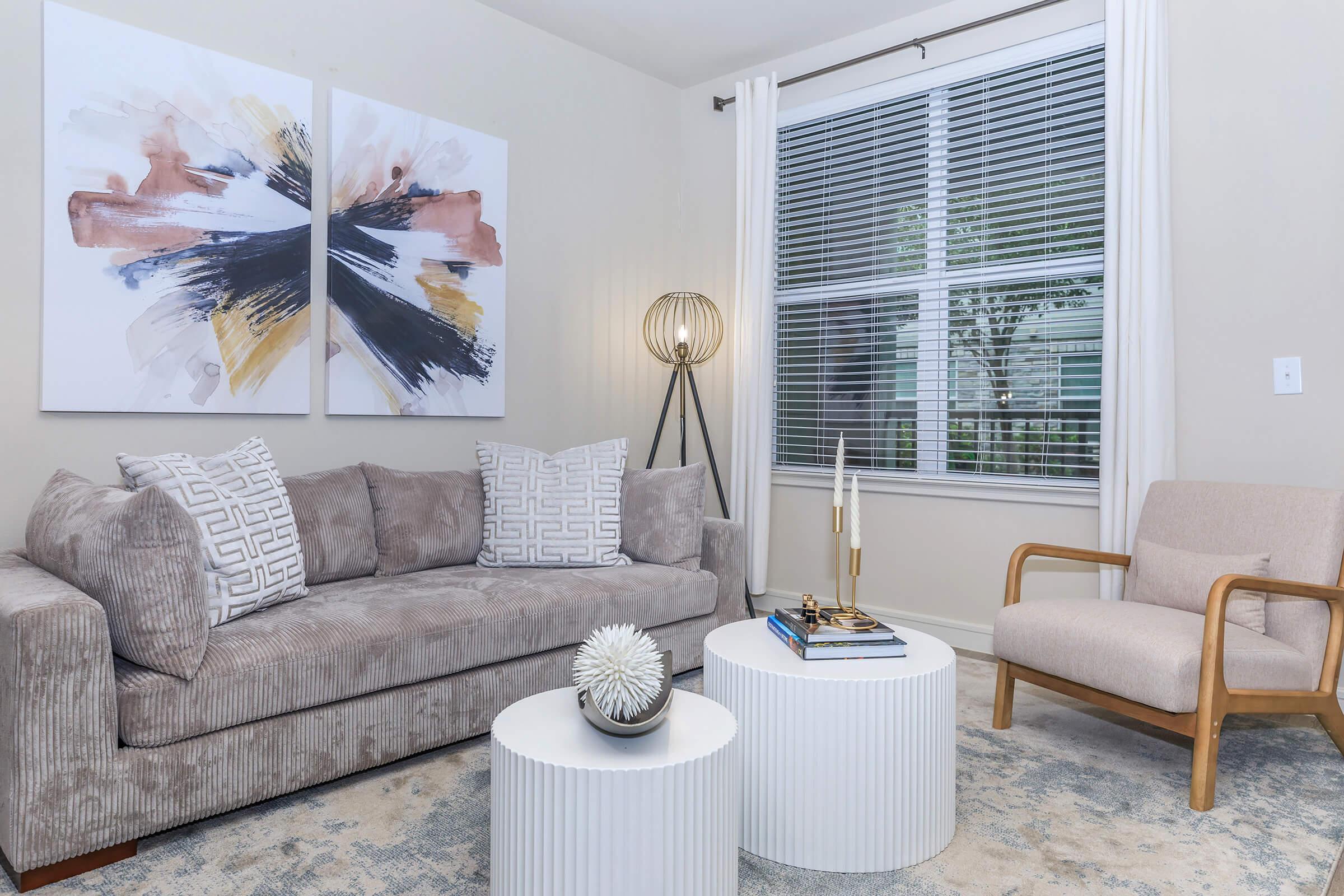 A cozy living room featuring a gray upholstered sofa with decorative pillows, two round white coffee tables, and a modern armchair. There's a floor lamp and abstract wall art with shades of black, white, and pink. Natural light enters through a window with white curtains, enhancing the inviting atmosphere.