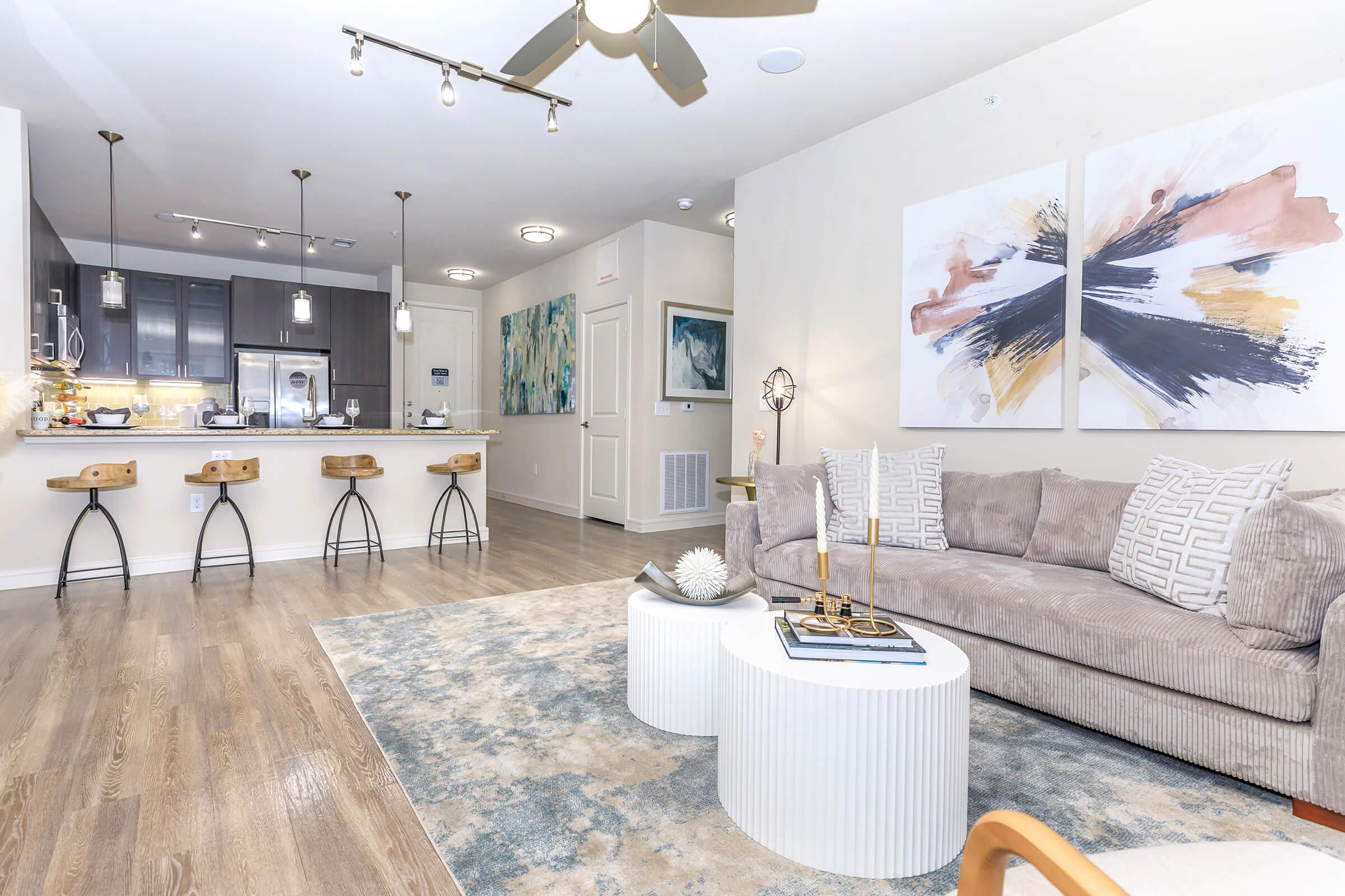 A modern living space featuring a light-colored sofa with decorative pillows, a round coffee table, and a large area rug. In the background, a kitchen with sleek cabinets and bar stools is visible. Abstract artwork adorns the wall, adding a touch of color to the neutral decor. Bright lighting enhances the open layout.