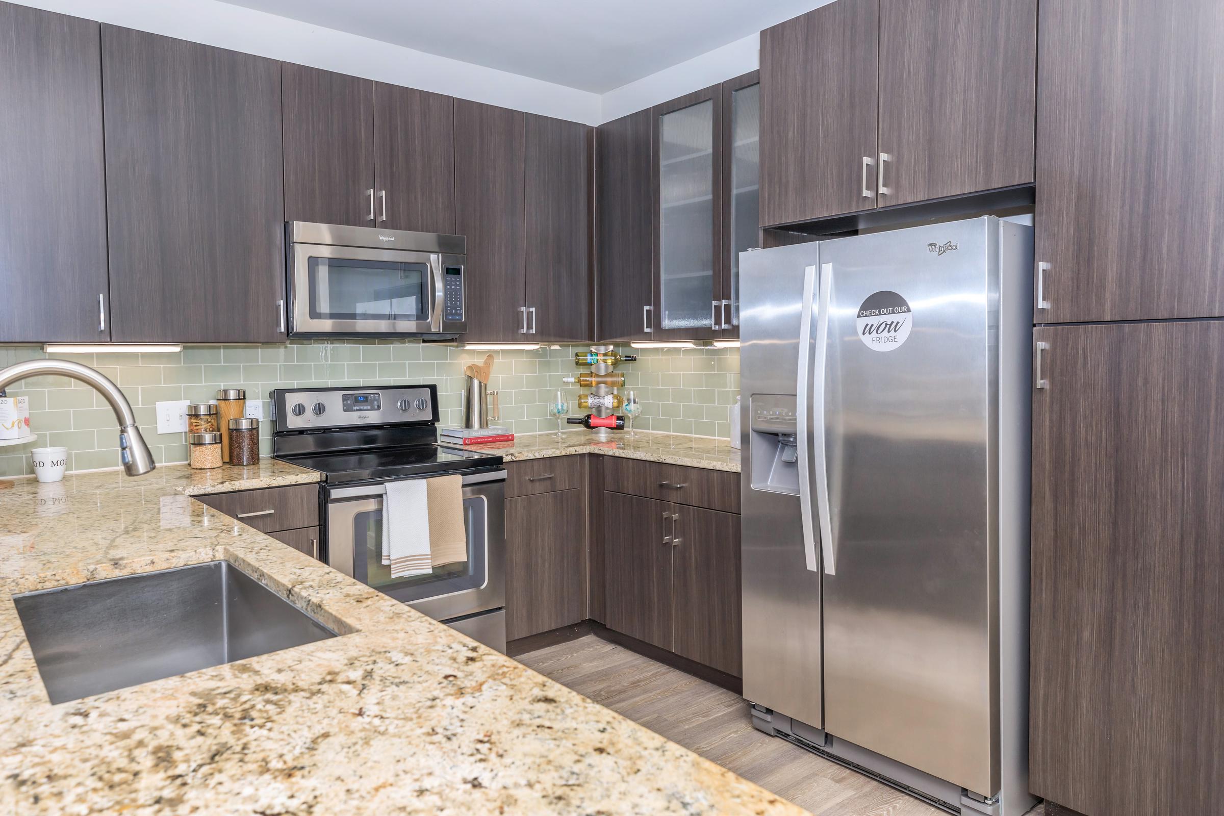 Modern kitchen with dark wood cabinetry, stainless steel appliances including a microwave, oven, and refrigerator. The countertop is made of granite, and the backsplash features pale green tiles. A sink with a modern faucet is visible, along with kitchen accessories and a small display of items on a shelf.