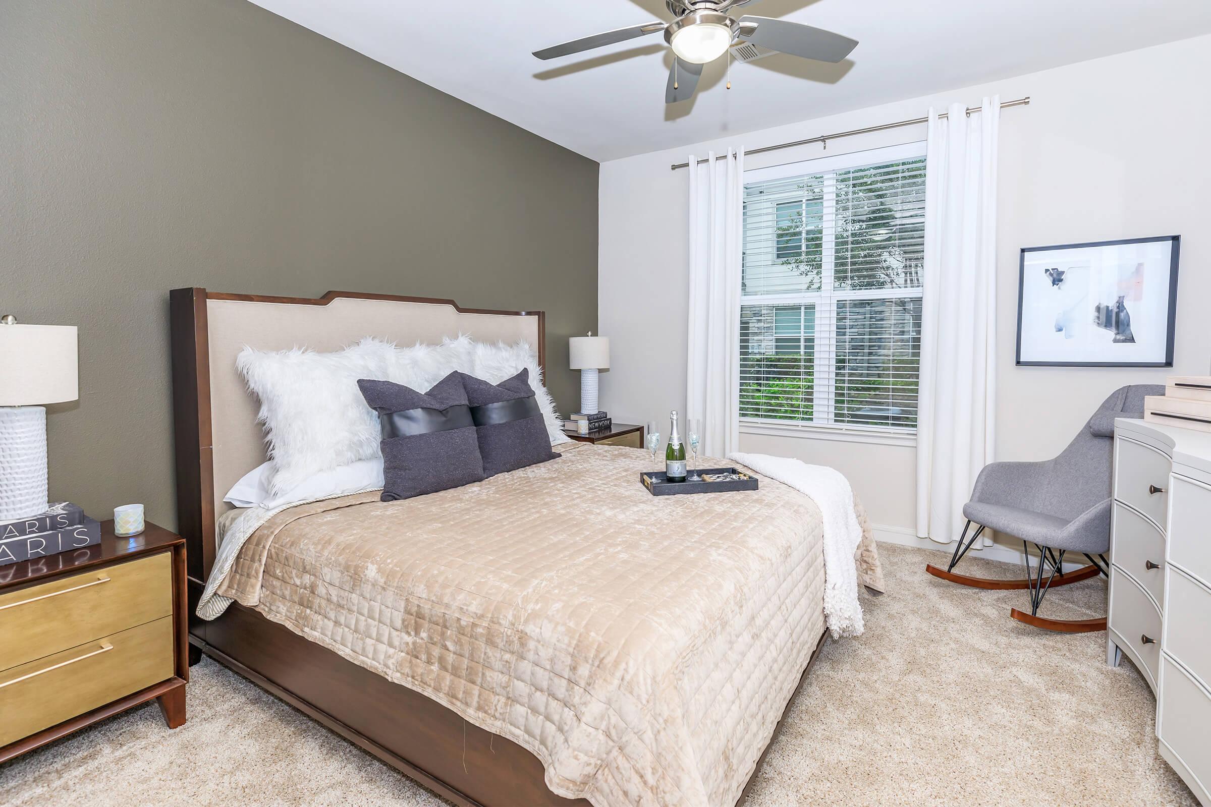A cozy bedroom featuring a comfortable bed with decorative pillows, a neutral-colored quilt, and a stylish headboard. There are two bedside lamps on wooden nightstands, a modern chair in the corner, and a window with sheer curtains allowing natural light. A small artwork hangs on the wall beside the window.