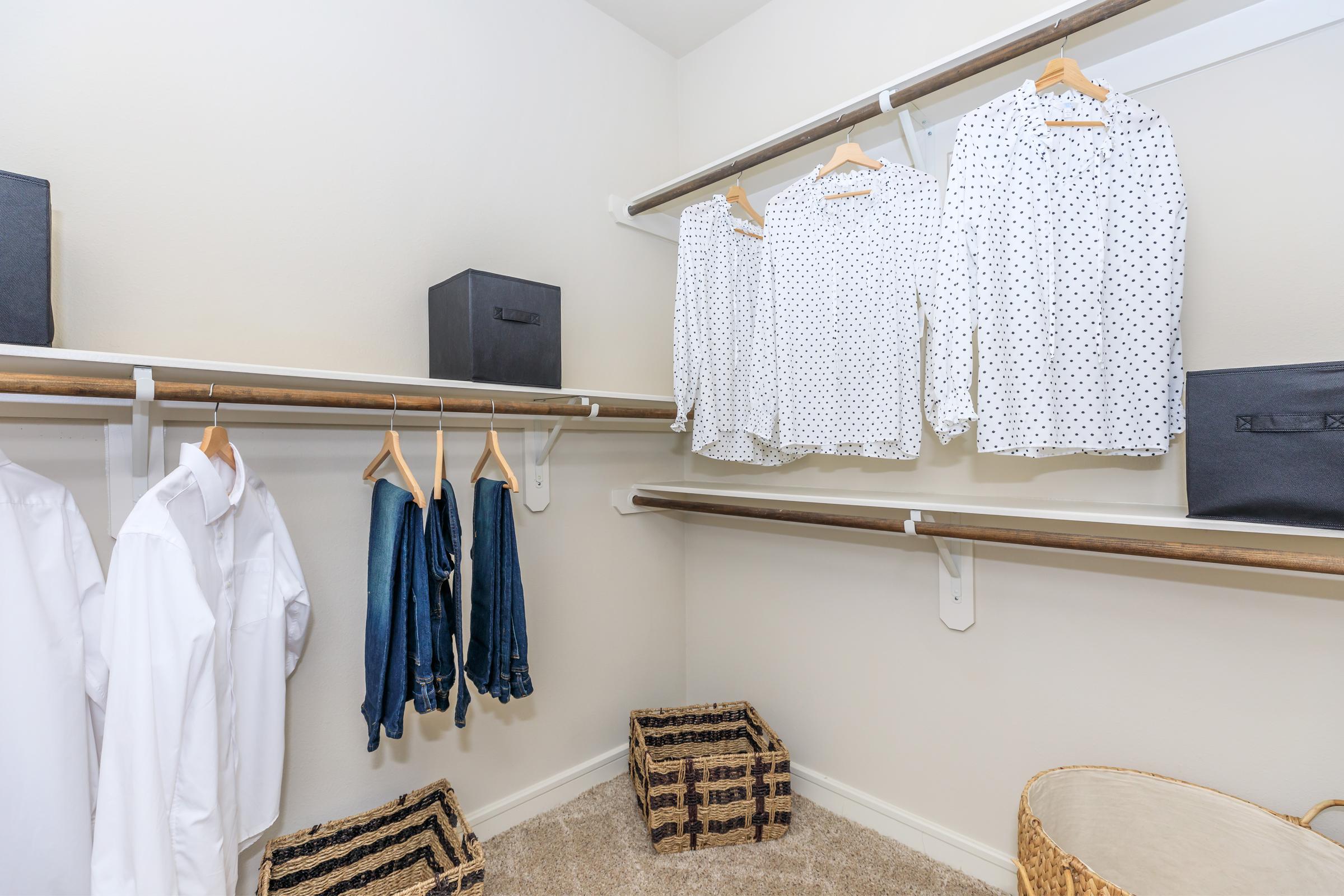 A well-organized closet featuring hanging white shirts with polka dots, denim jeans, and storage boxes. There are also woven baskets on the floor, highlighting a neat and tidy appearance. The walls are a neutral color, and the overall ambiance is bright and spacious.