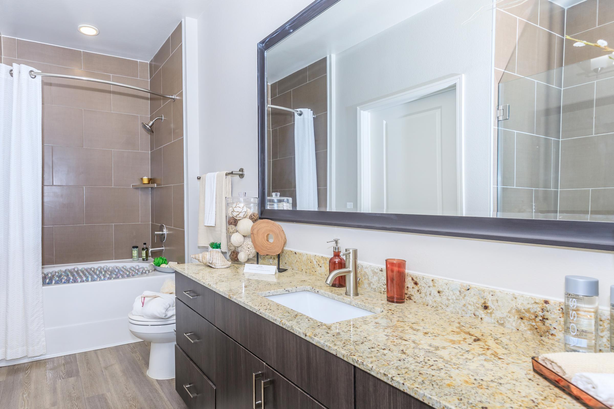 A modern bathroom featuring a light granite countertop with a sink, dark wood cabinetry, and a large mirror. The bathtub is partially visible, with a shower curtain and decorative items on the countertop, including a soap dispenser and candles. The walls are tiled in neutral tones.