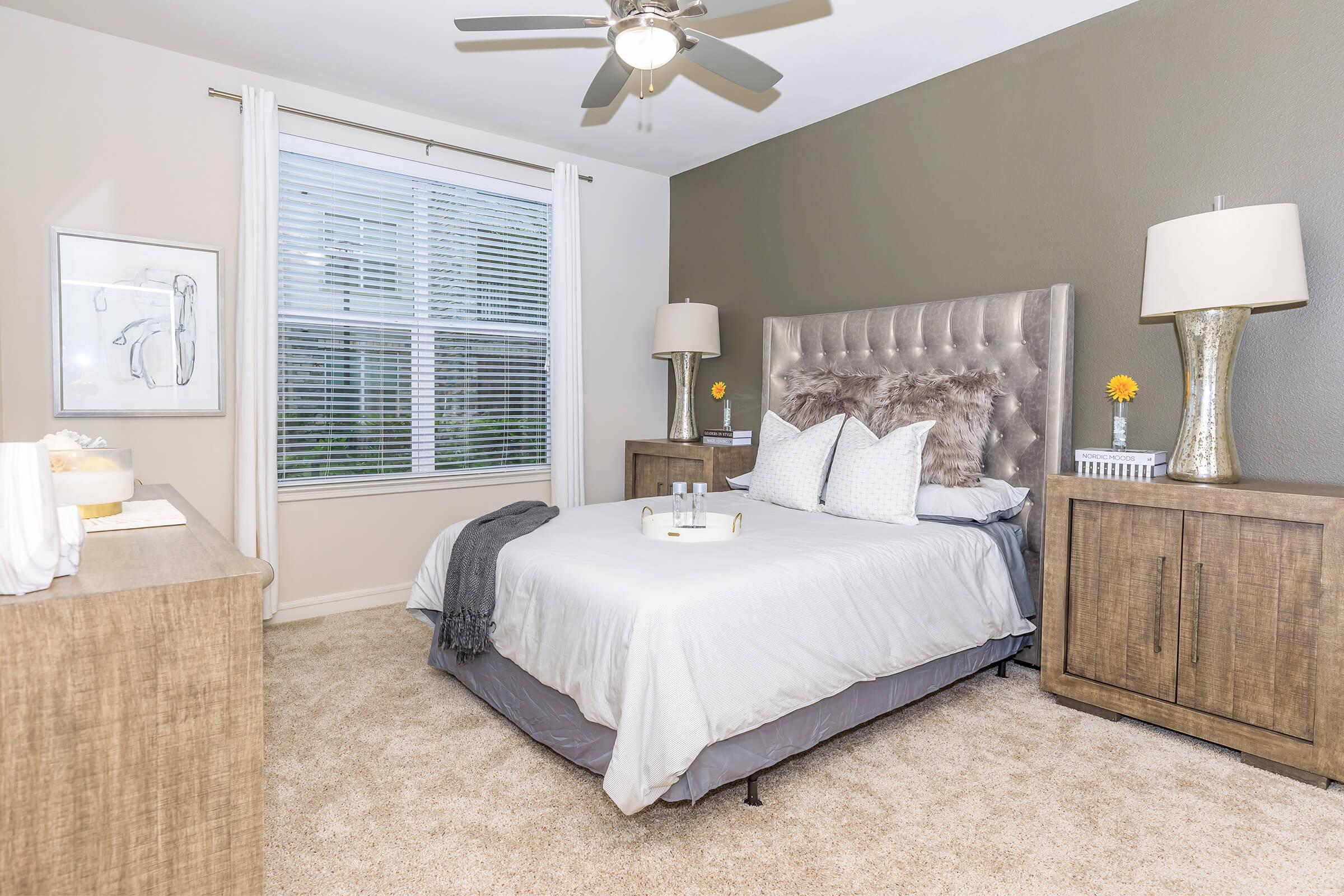A cozy, well-decorated bedroom featuring a plush bed with gray bedding and decorative pillows. A stylish headboard is complemented by two bedside lamps and a small table with a flower vase. Light-colored walls and a window with sheer curtains enhance the bright, inviting atmosphere. Beige carpet adds warmth to the room.