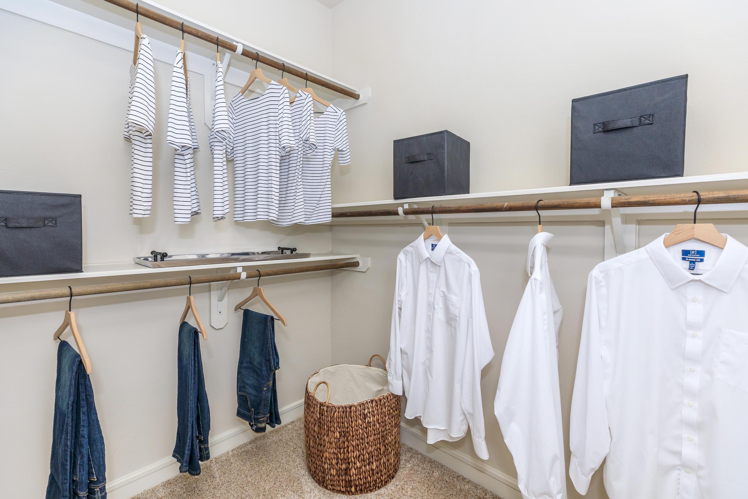 A neatly organized closet featuring striped shirts hanging on hangers, white dress shirts, and denim jeans. Two black storage boxes are placed on the shelves, and a round woven basket is positioned on the floor, contributing to the tidy and minimalist aesthetic.