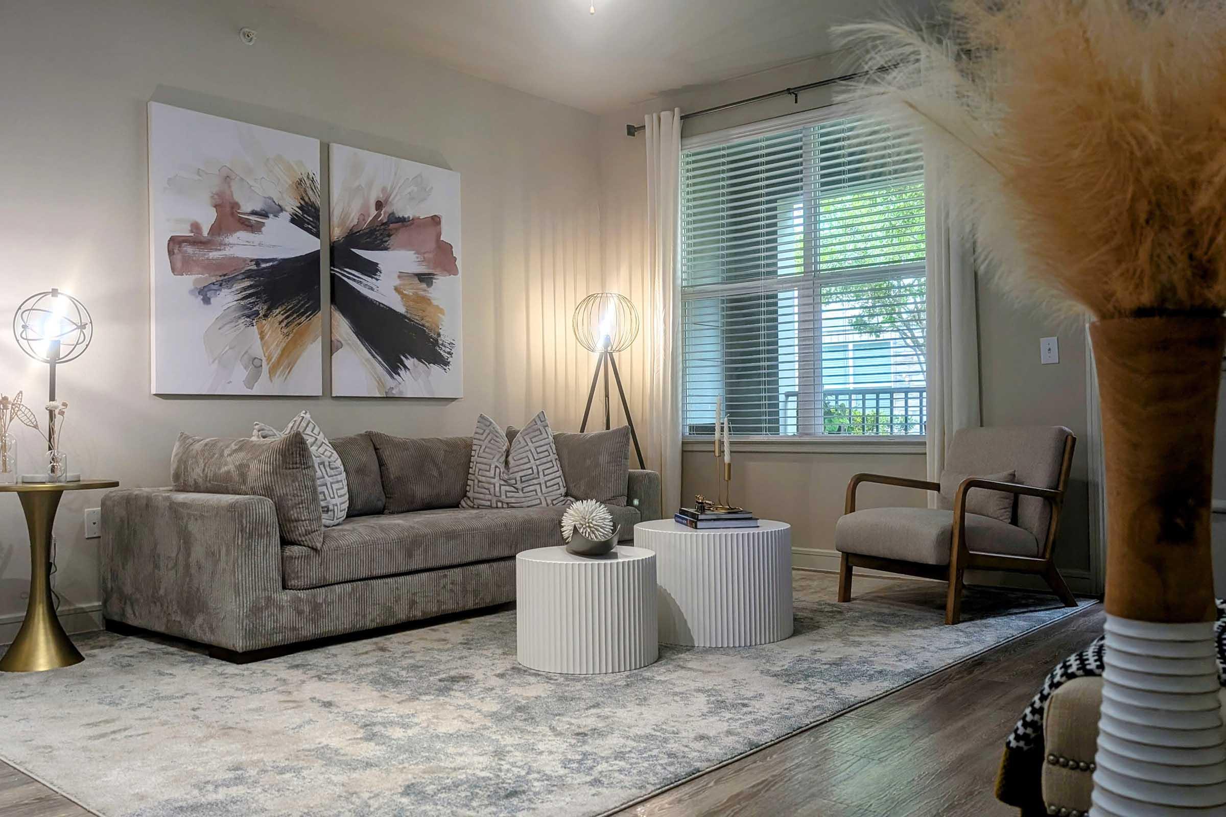 A cozy living room featuring a gray sofa with patterned cushions, two abstract art pieces on the wall, a round coffee table, and a stylish armchair. Soft lighting enhances the inviting atmosphere, with a decorative plant and neutral colors throughout the space. Large window lets in natural light.