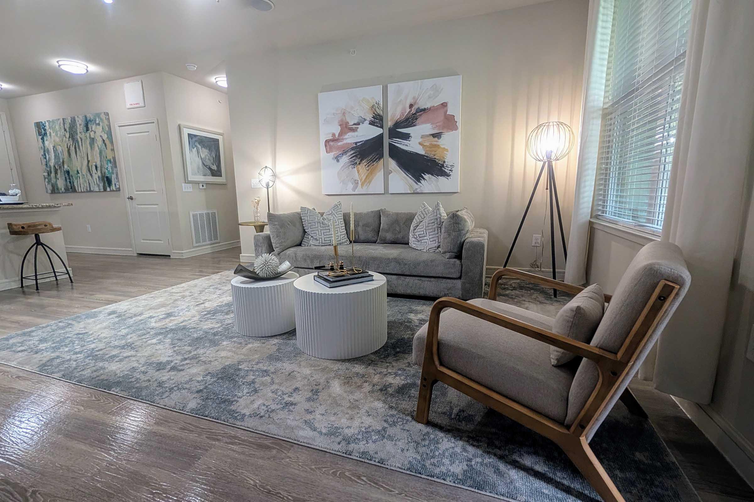 A modern living room featuring a gray sofa with decorative pillows, two round white coffee tables, and a stylish floor lamp. The decor includes abstract artwork on the walls and natural light streaming in through large windows. A cozy armchair complements the seating area, situated on a soft area rug.