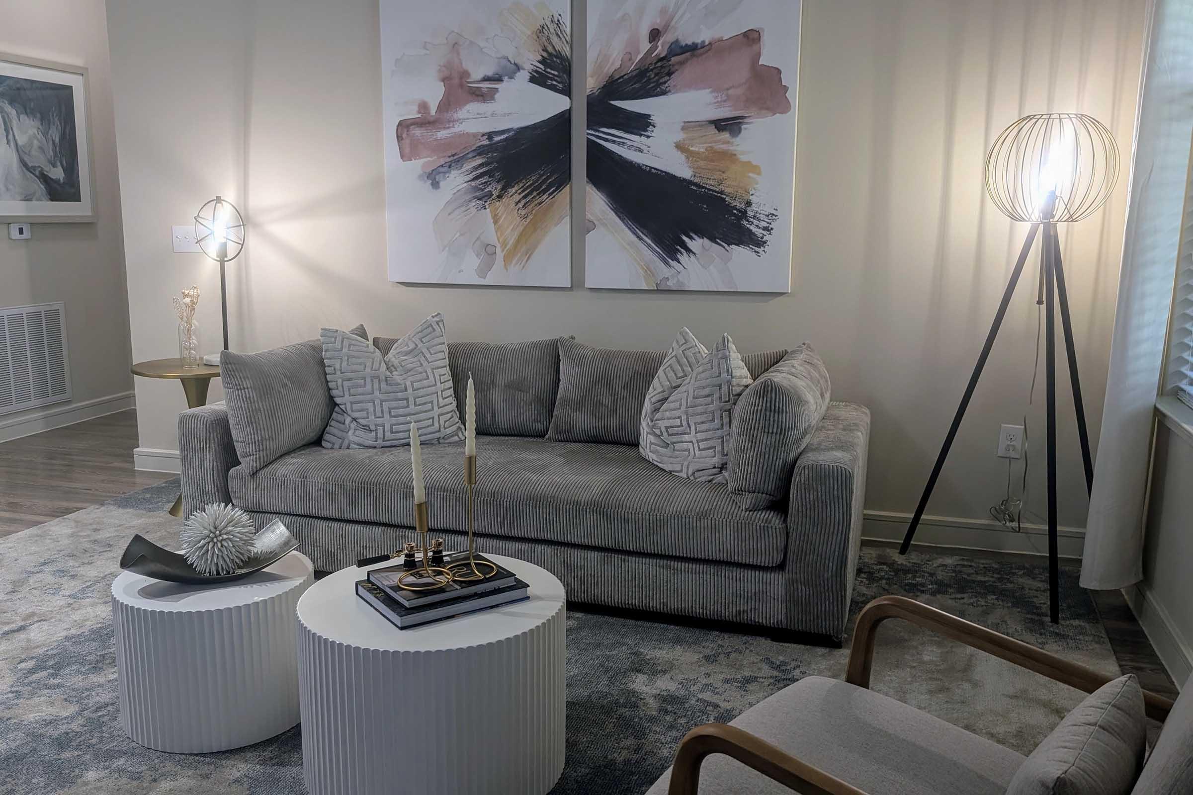 A cozy living room featuring a gray sofa adorned with patterned pillows, two white side tables with decorative items, and a stylish floor lamp. Abstract art hangs on the wall, adding a modern touch to the space, which is illuminated by natural light from a window.