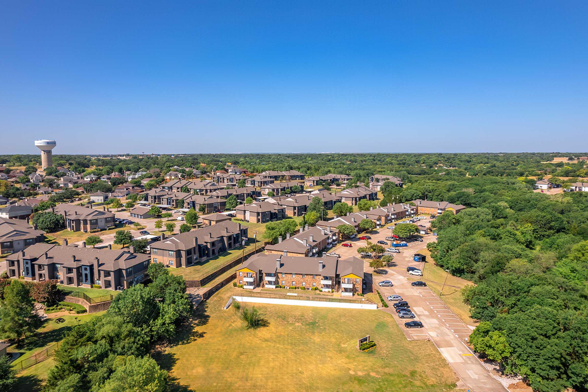 Aerial view of a suburban neighborhood featuring multiple residential buildings, parking areas, and green spaces, with a water tower visible in the distance and clear blue skies overhead. The scene captures a blend of housing and nature, showcasing well-maintained properties and organized streets.