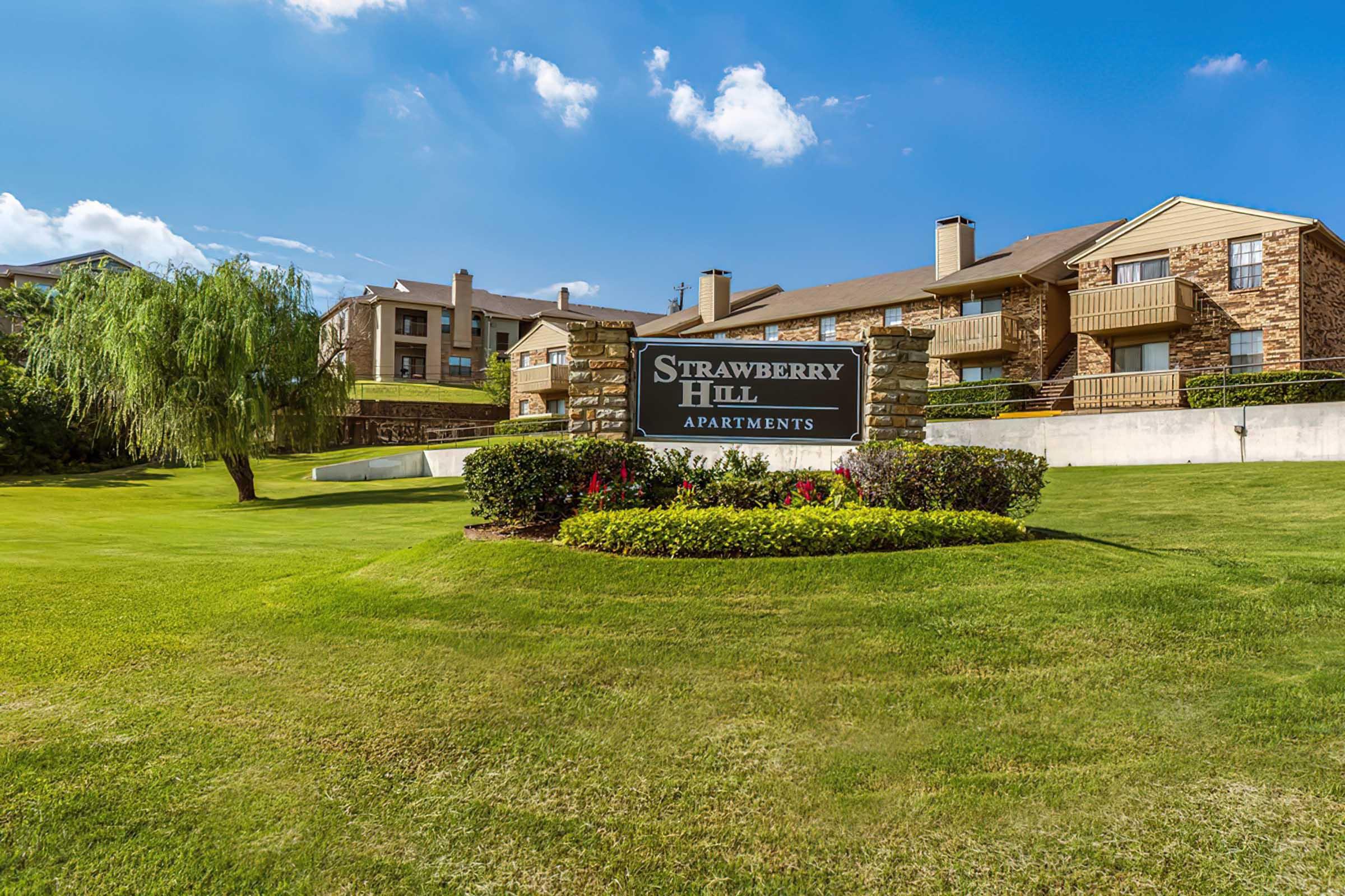 A scenic view of the Strawberry Hill Apartments sign, surrounded by well-maintained green lawns and colorful flower beds. In the background, there are two buildings displaying a mix of brick and beige siding under a clear blue sky with fluffy white clouds.