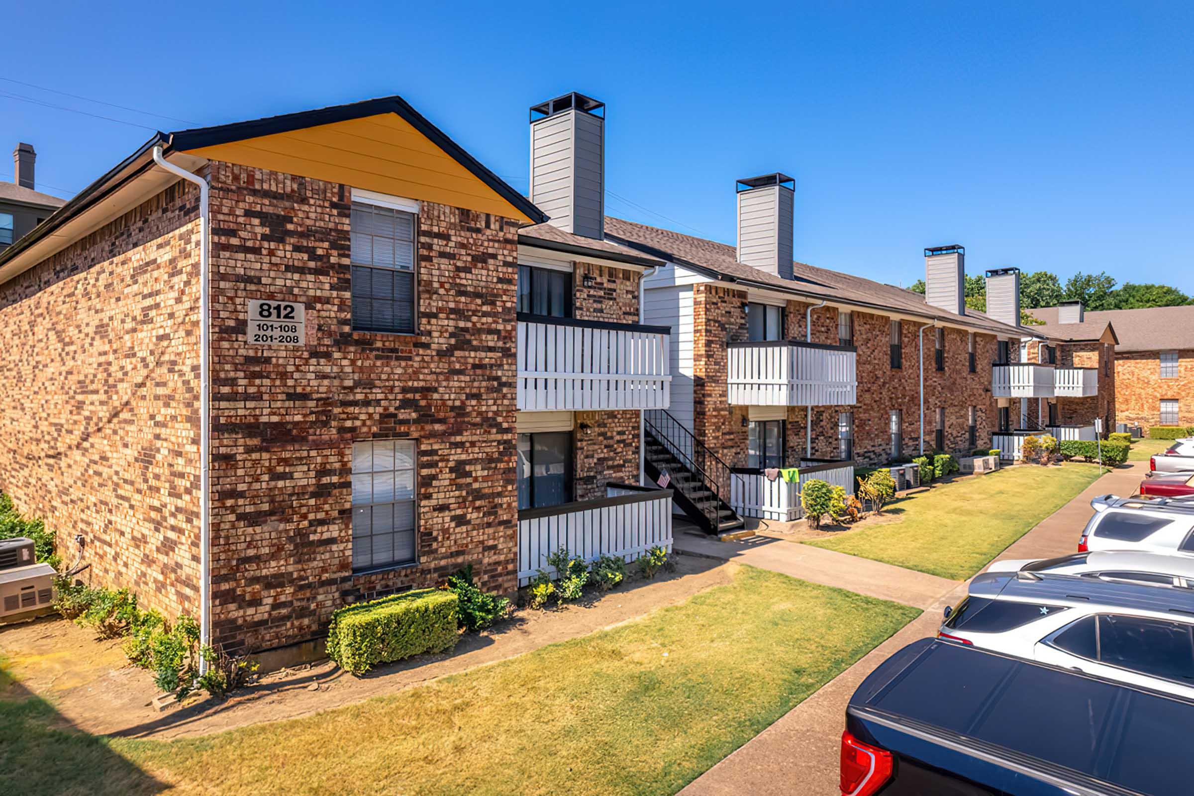 Two-story brick apartment building with balconies and a yellow awning. Green lawn and parking lot with several cars in the foreground. Clear blue sky provides a bright backdrop. Property signage visible on the building exterior.