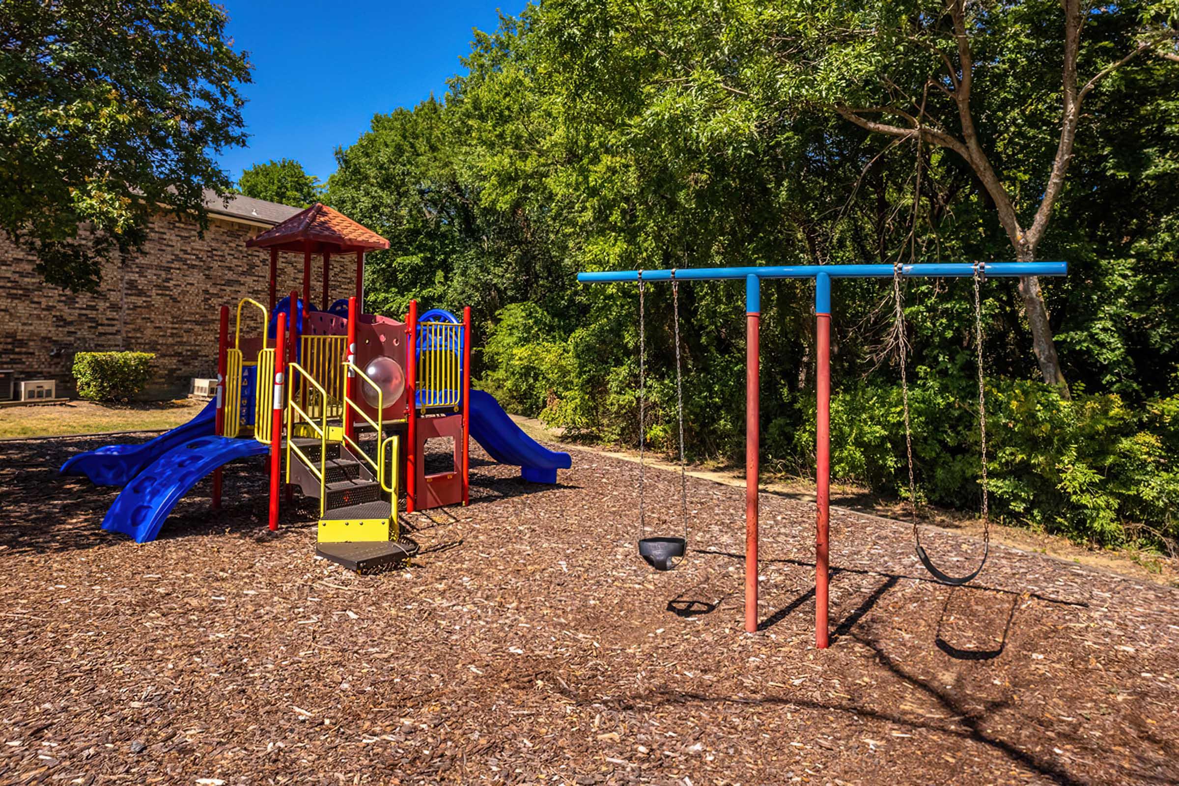 Colorful playground equipment in a sunny outdoor setting, featuring a slide, climbing structure, and swings. Surrounding greenery and mulch ground create a inviting atmosphere for children to play.