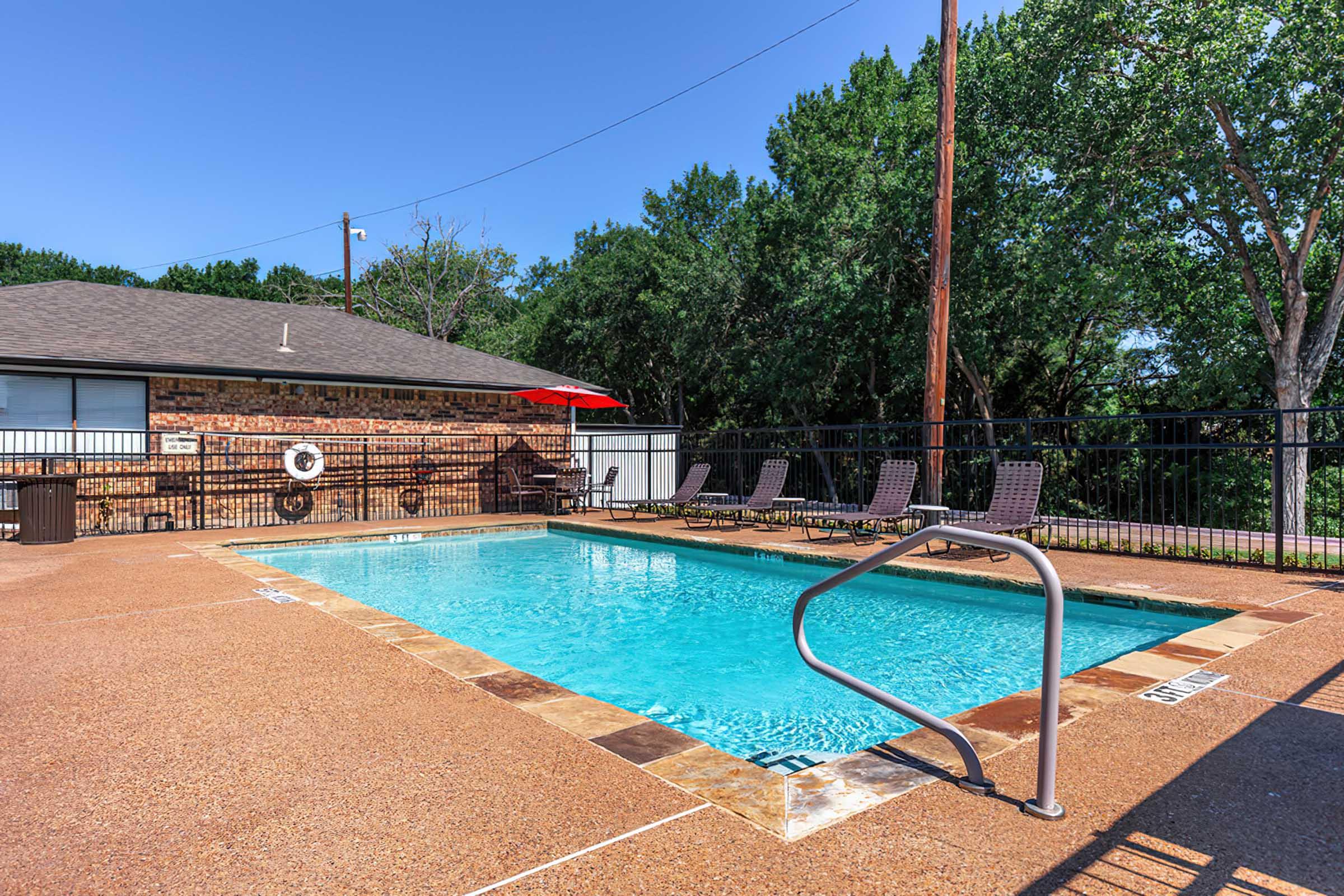 A clear blue swimming pool surrounded by a tan concrete deck, with several lounge chairs arranged neatly nearby. The background features a brick building and green trees under a bright blue sky. An umbrella provides shade over part of the pool area.