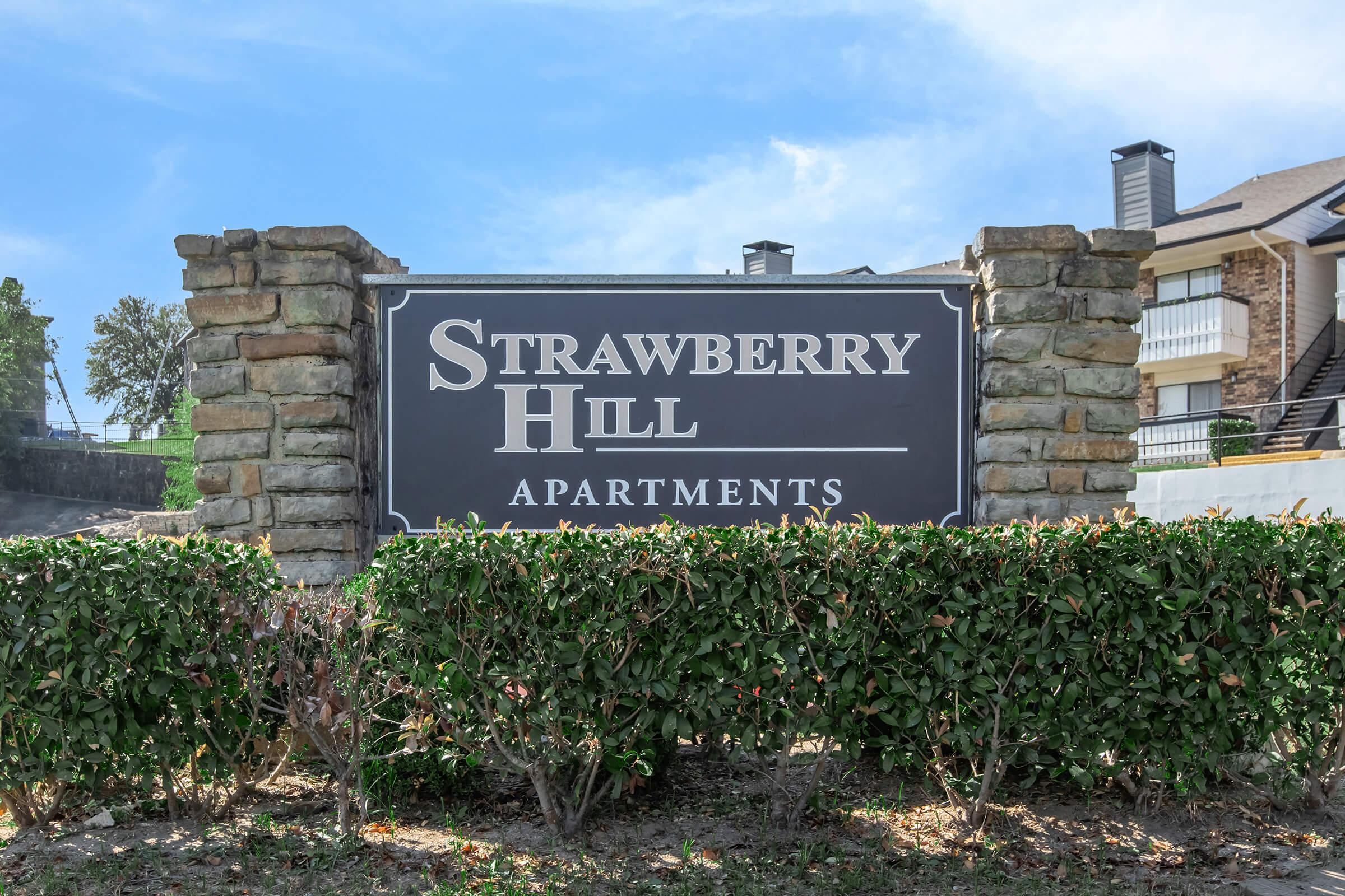 Sign for Strawberry Hill Apartments, featuring a dark background with white text. The sign is framed by stone columns and surrounded by bushes against a clear blue sky.