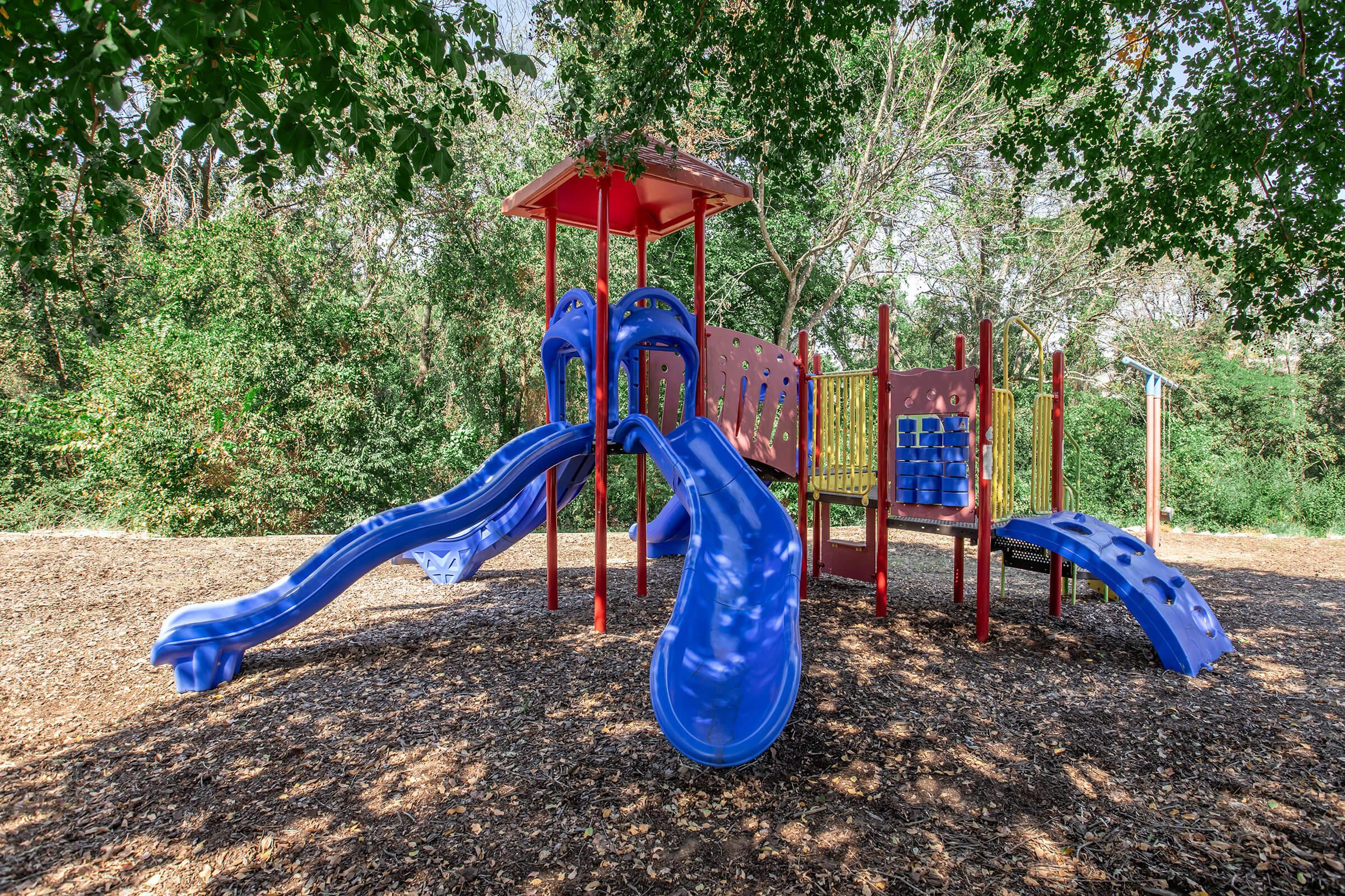 A brightly colored playground structure featuring two blue slides, climbing areas, and a playhouse, surrounded by trees and natural greenery. The ground is covered with wood chips, providing a soft play surface for children.