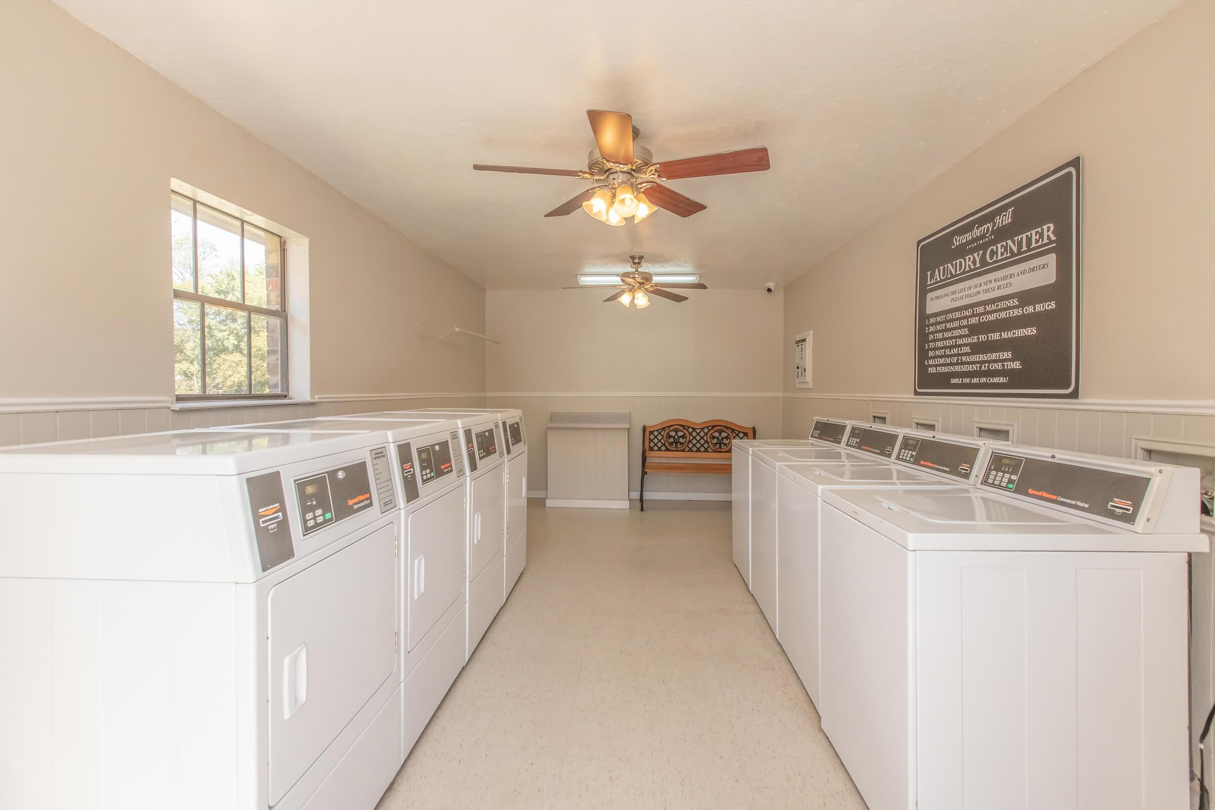 A clean, well-lit laundry room featuring several white washing machines lined up against the wall. Two ceiling fans are visible, and there’s a bench for seating. A sign on the wall reads "Laundry Center." Large windows allow natural light to enter the space.