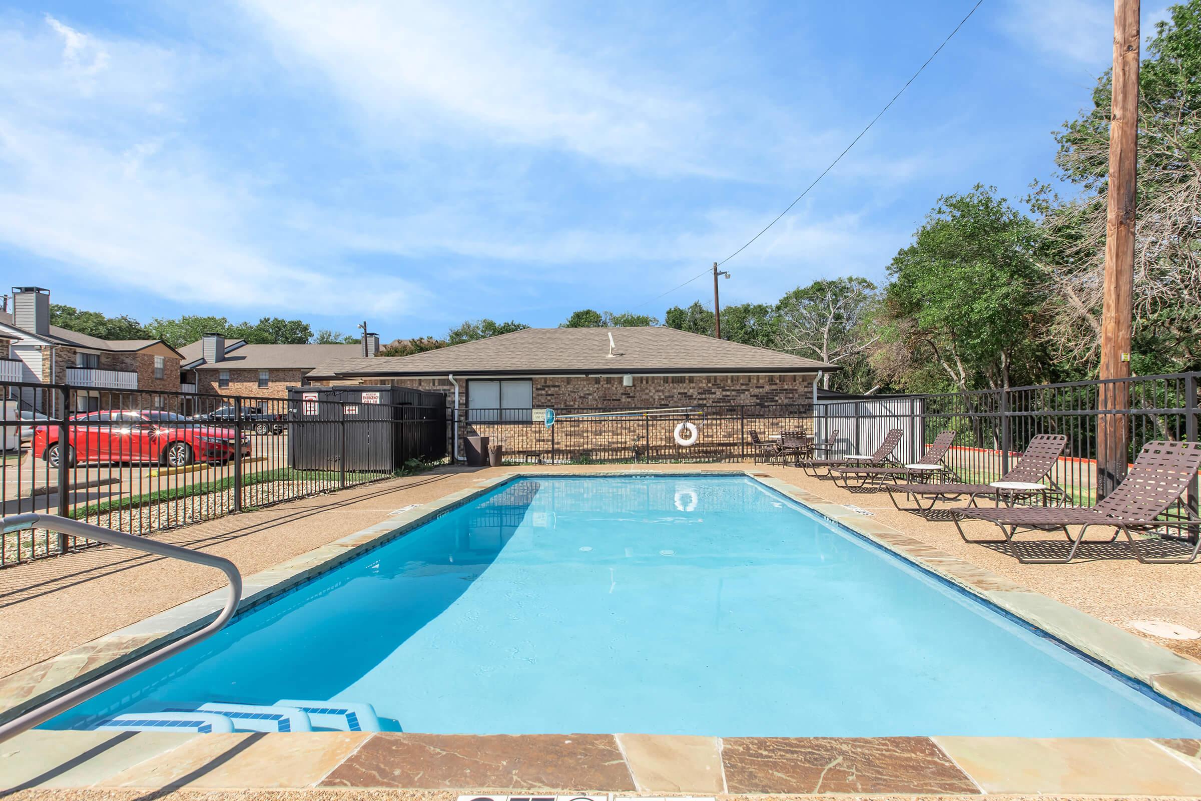 A clear swimming pool with bright blue water surrounded by a fence. Lounge chairs are positioned by the poolside. In the background, a single-story building can be seen among trees under a clear blue sky.