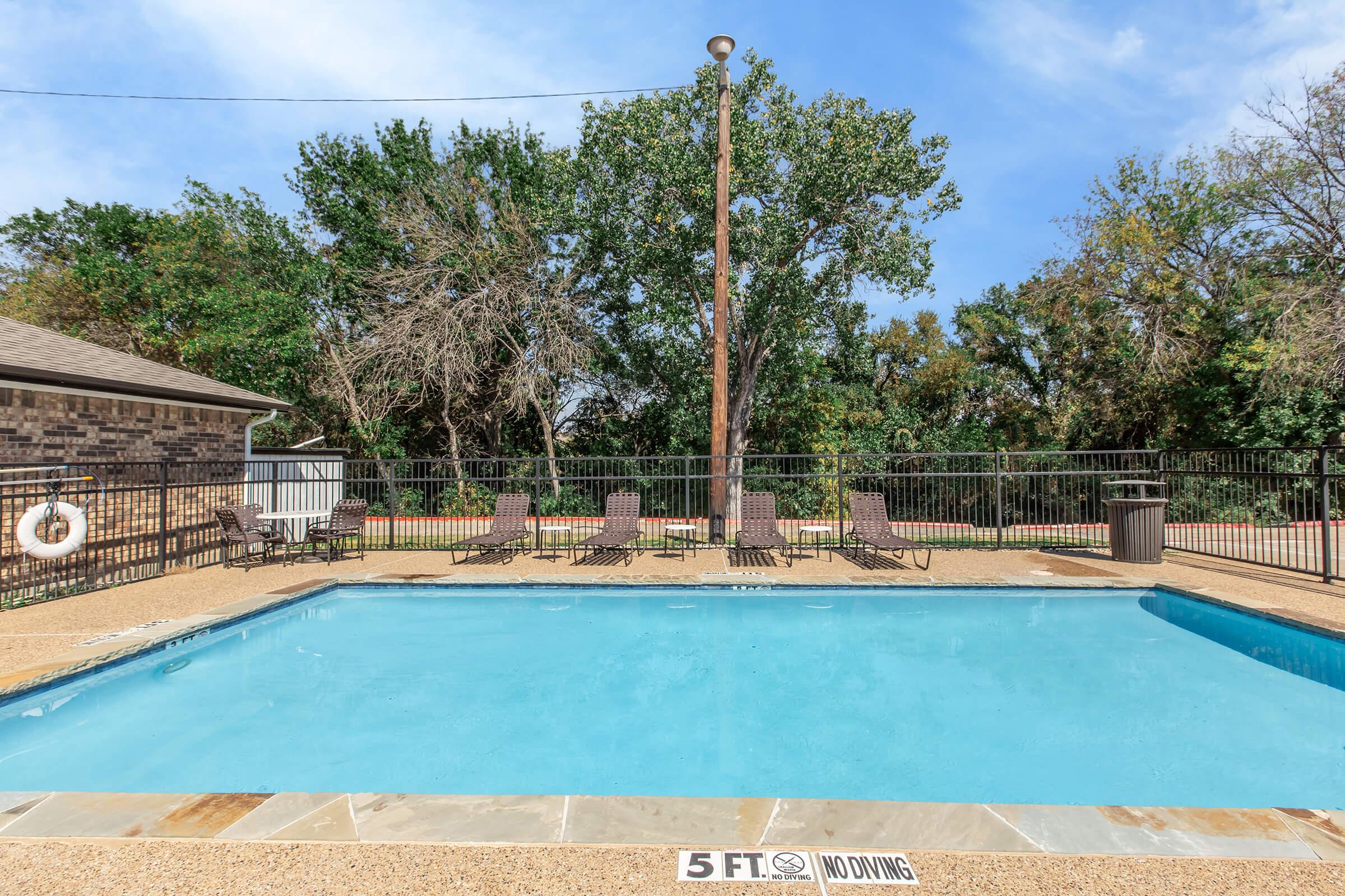 Swimming pool surrounded by a concrete deck, with several lounge chairs placed around it. A wooden fence encloses the area, and trees can be seen in the background. A sign indicates a depth of 5 feet and a "No Diving" rule. Bright blue sky partially visible above.
