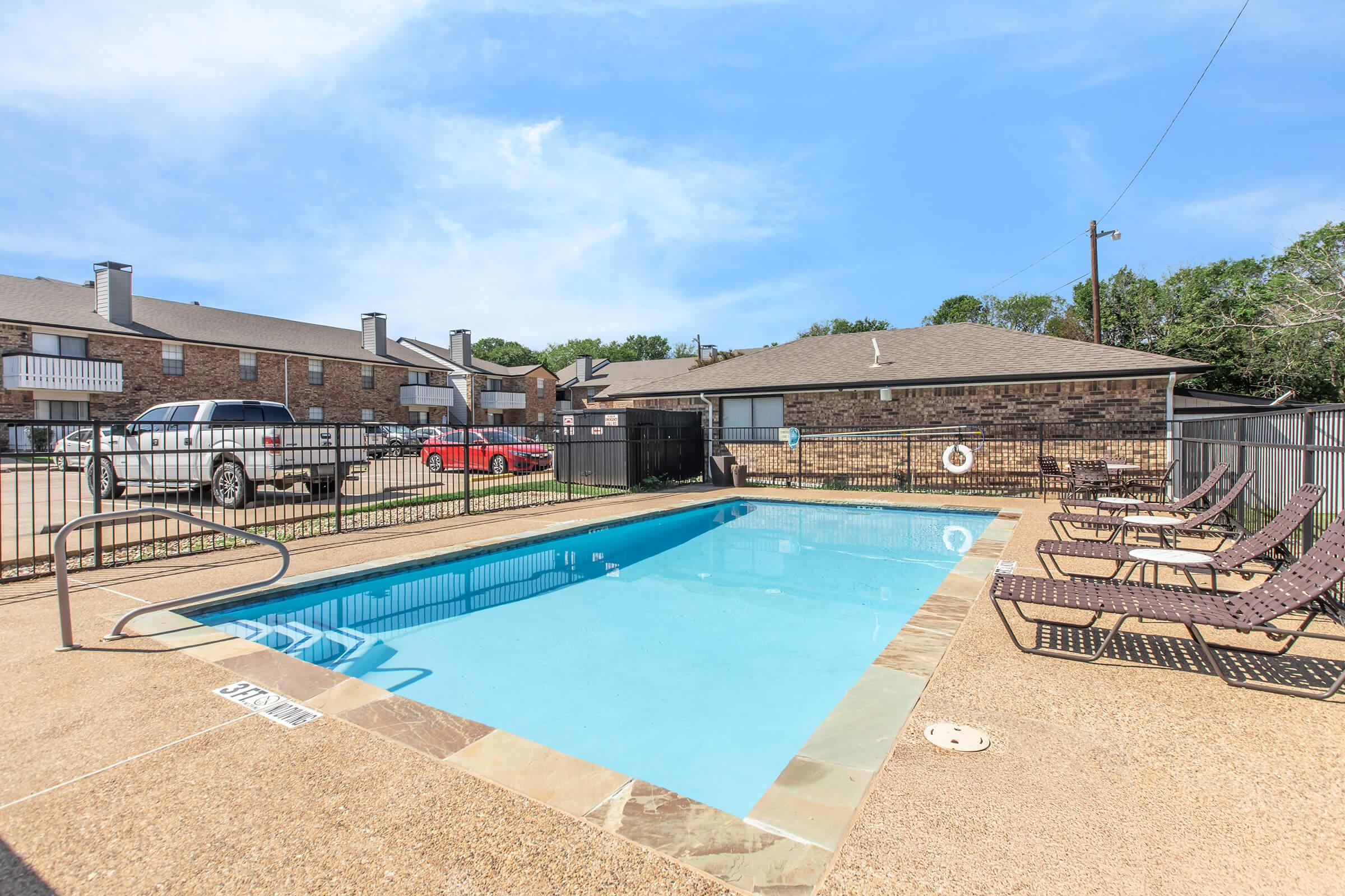 A clear blue swimming pool surrounded by a gated area. Lounge chairs are placed along one side of the pool, and two cars are parked nearby. There are residential buildings in the background under a bright blue sky.