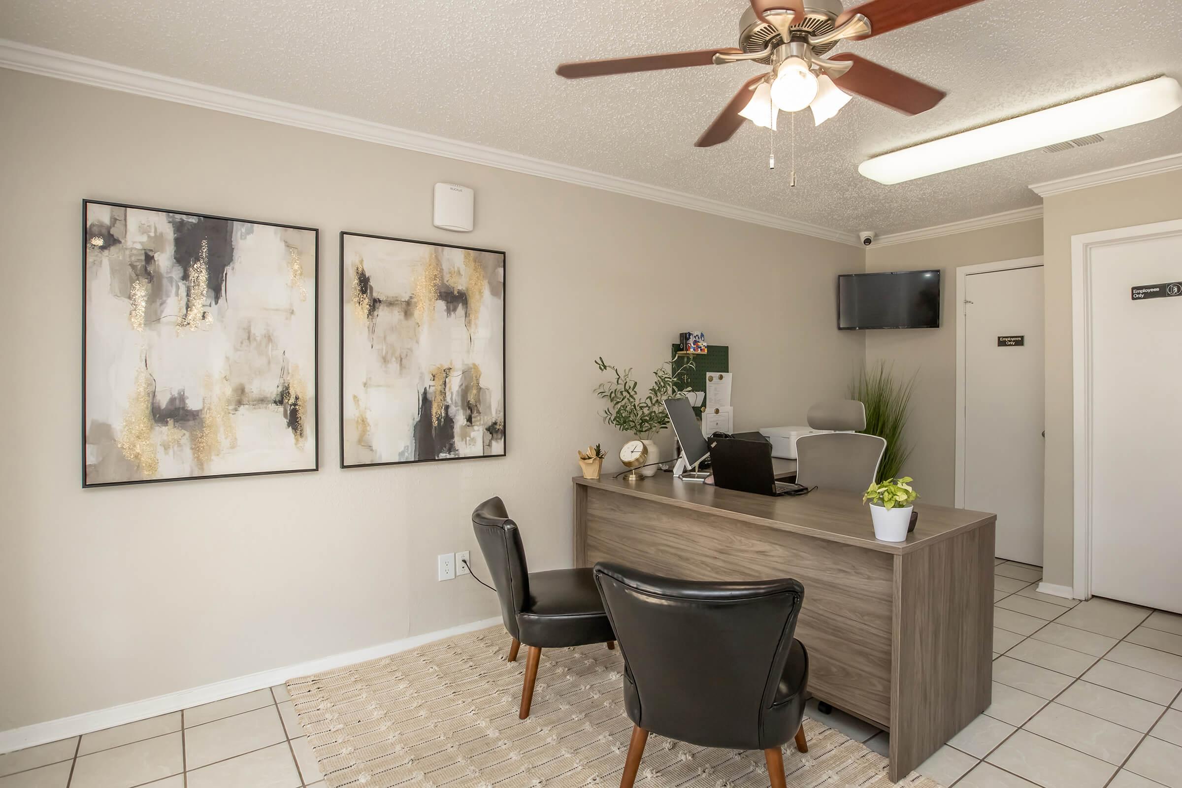 A modern office reception area featuring a sleek wooden desk with a computer and plant, two black chairs facing the desk, and abstract wall art on either side. A ceiling fan and a TV are visible, along with light-colored walls and tiled flooring, creating a welcoming professional environment.