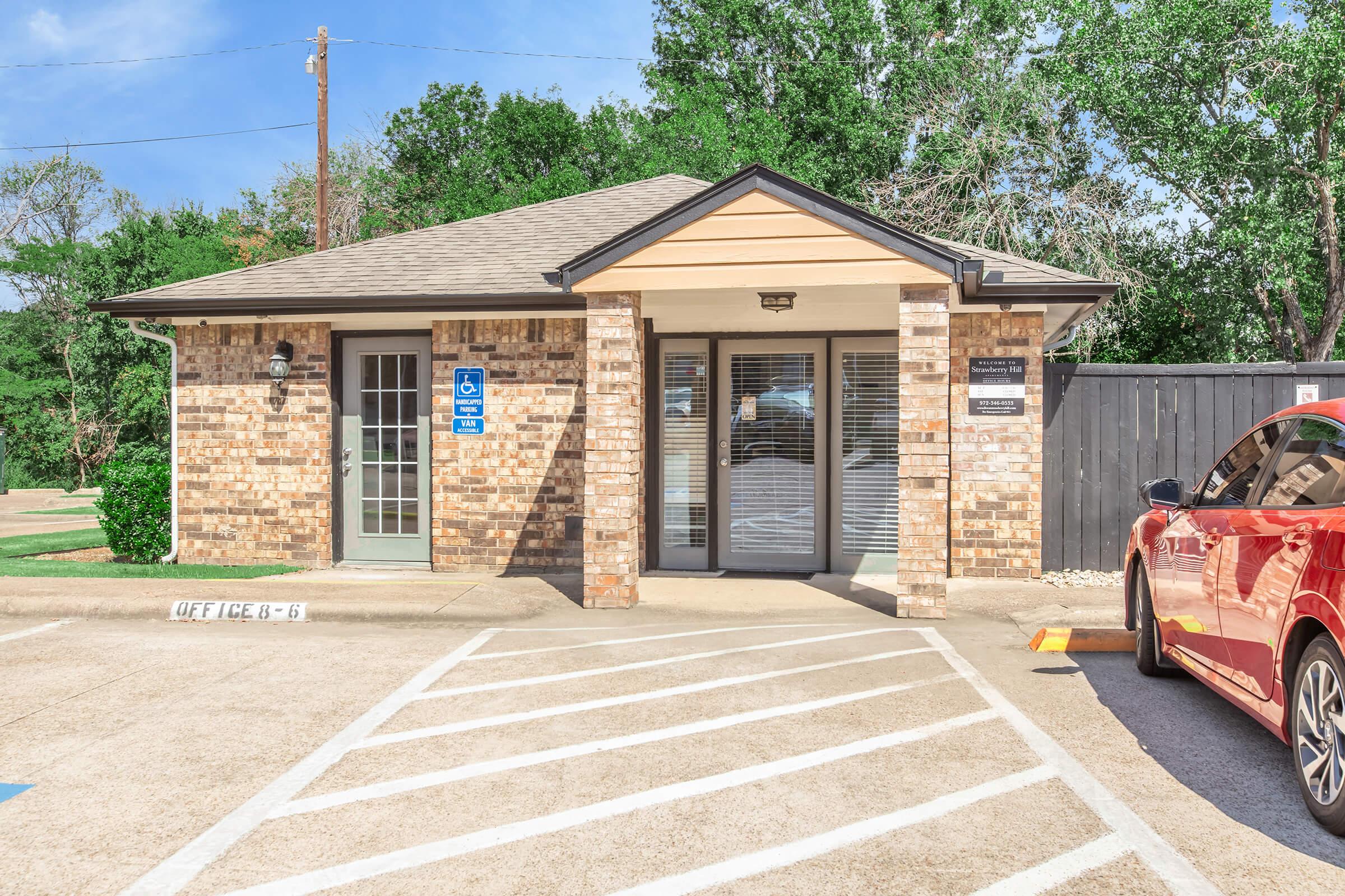 Single-story brick building with a peaked roof and large windows. There is a sign indicating "Office" on the front. A red car is parked in a designated spot nearby. The surrounding area features green trees and a clear blue sky. Accessible parking spaces are visible in front of the building.
