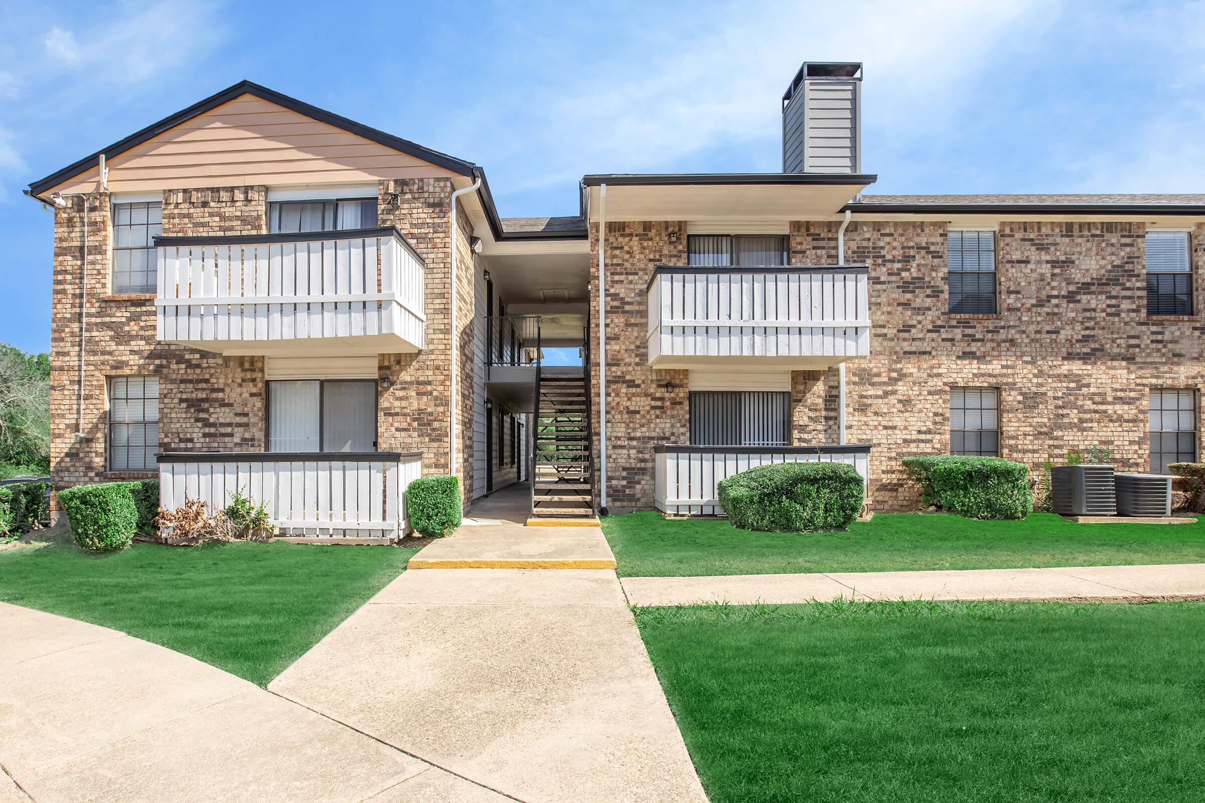 Two-story brick apartment building with several balconies. The building features a light-colored trim, and there are landscaped green areas with shrubs. A concrete pathway leads to the entrance, and the sky is clear with some scattered clouds.
