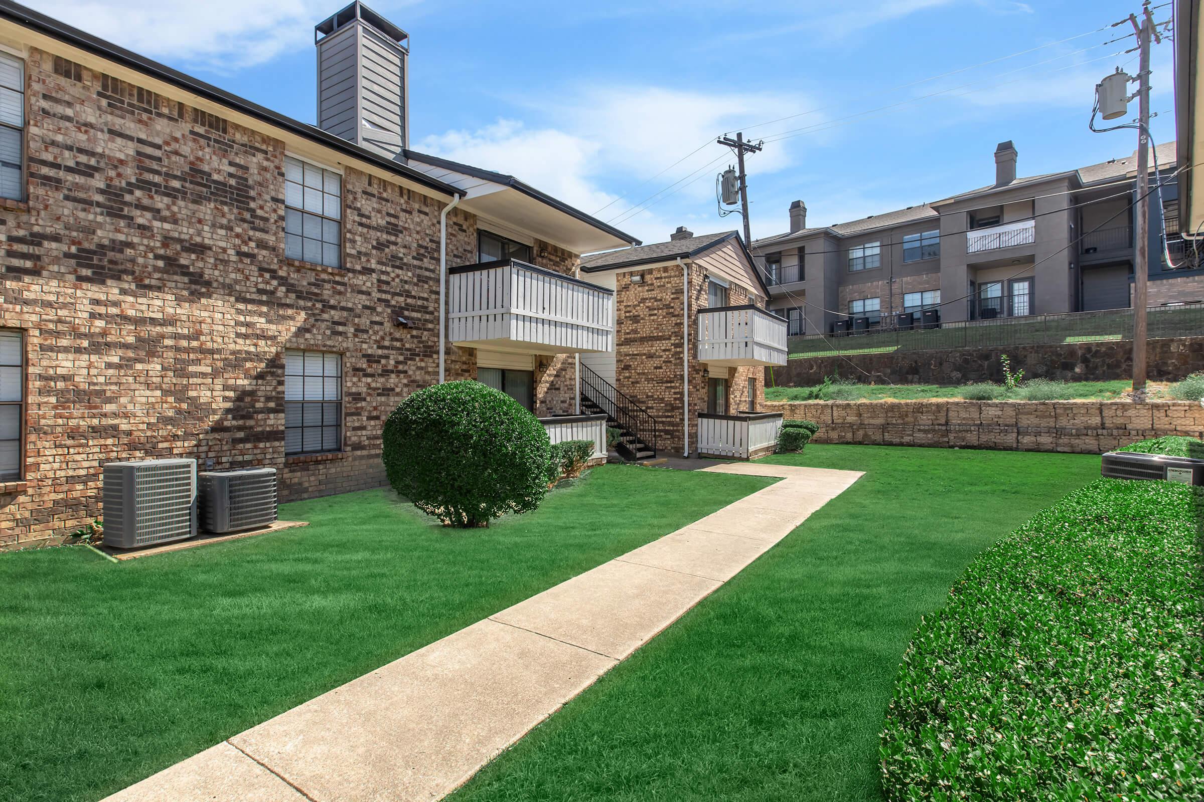 A well-maintained apartment complex featuring brick exterior buildings, manicured lawns, and a concrete pathway. Air conditioning units are visible beside the buildings, and utility poles are in the background against a clear blue sky.