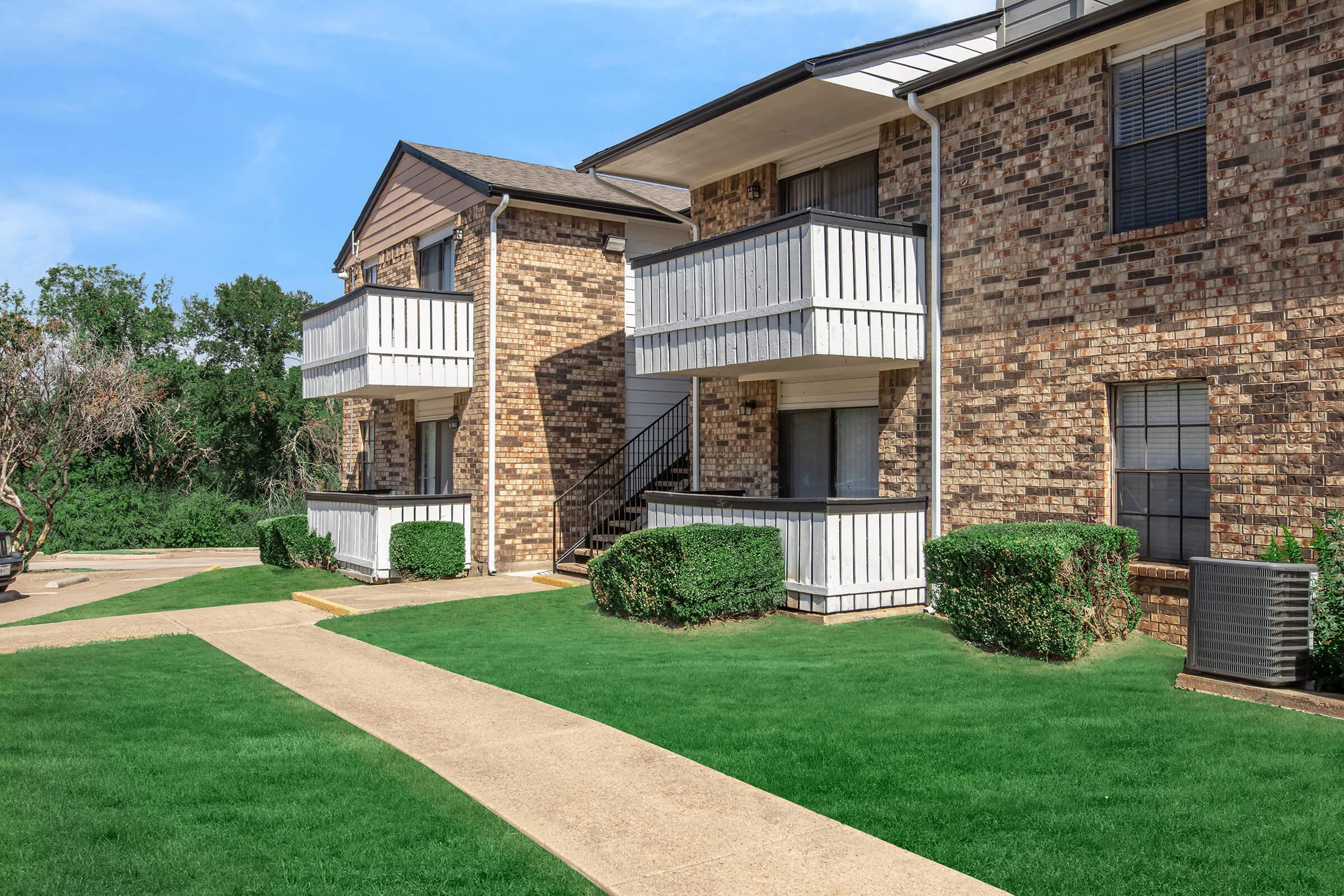 Two-story brick apartment building with white balconies, surrounded by neatly trimmed grass and small bushes. A concrete walkway leads to the entrance, with a clear blue sky in the background. The setting appears well-maintained and inviting.