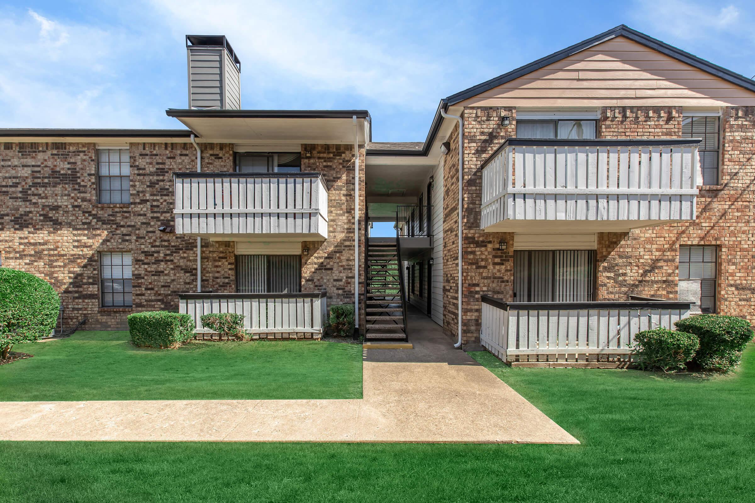 A view of an apartment building featuring two stories, with brick exterior and wooden balconies on either side. Lush green lawns and neatly trimmed hedges surround the building, and a walkway leads to the entrance, visible in the center. The sky is clear and bright.