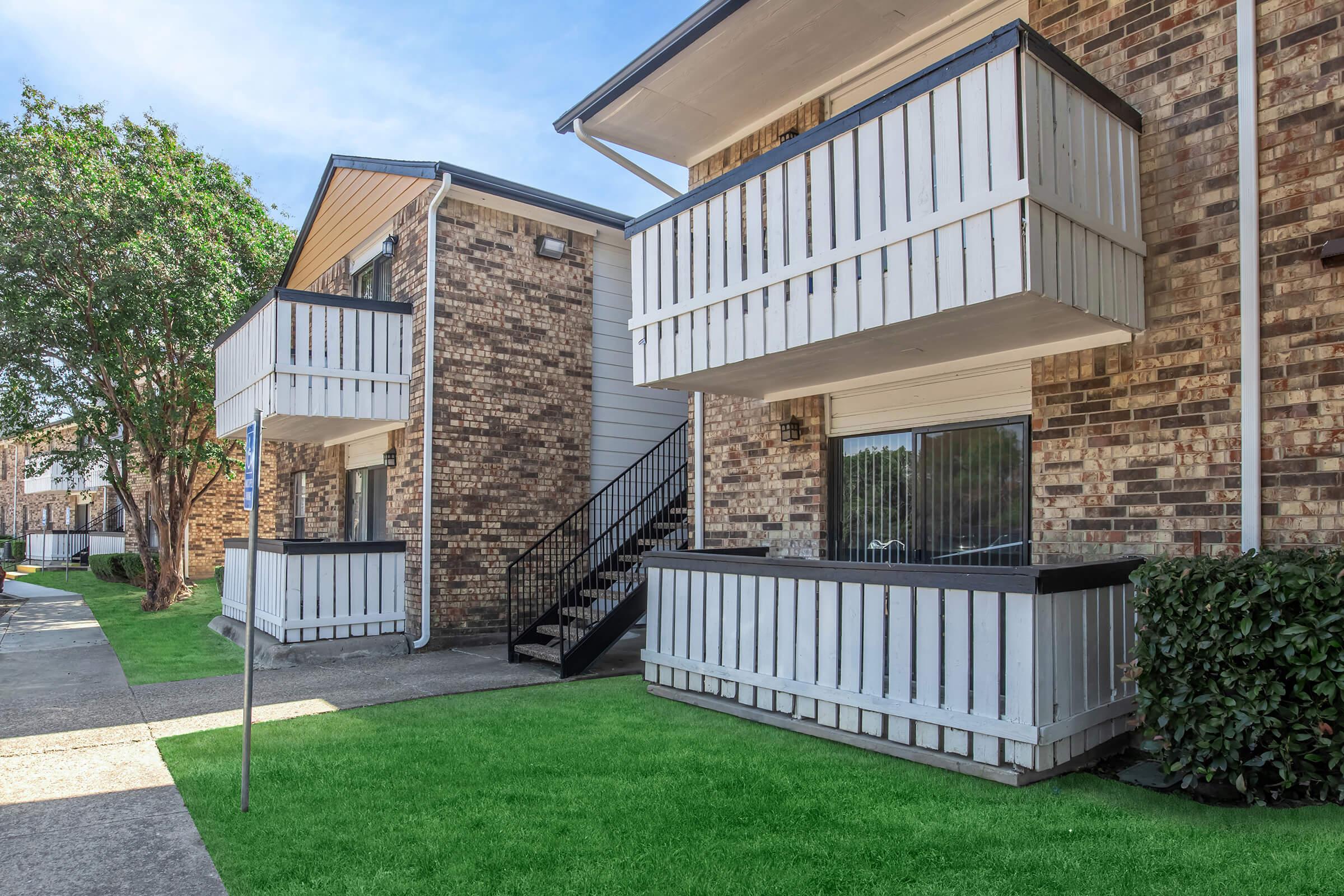 Two-story brick apartment building with white balconies. Each unit features a staircase leading to the upper level. The pathway is lined with green grass, and there are trees in the background under a clear blue sky.