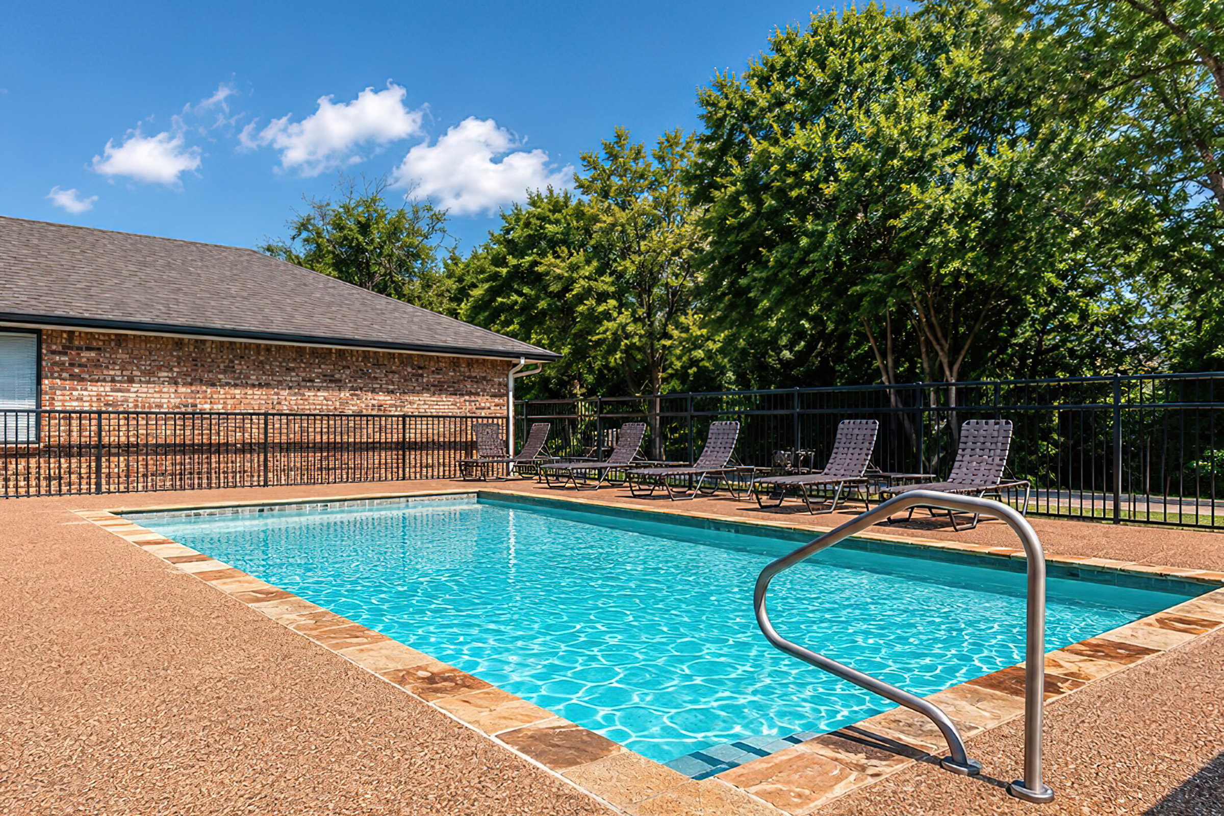 A refreshing swimming pool with clear blue water, surrounded by a tan concrete deck. Several lounge chairs are lined up nearby, with a fence providing privacy. The sky is bright blue with a few fluffy clouds, and green trees provide a natural backdrop.