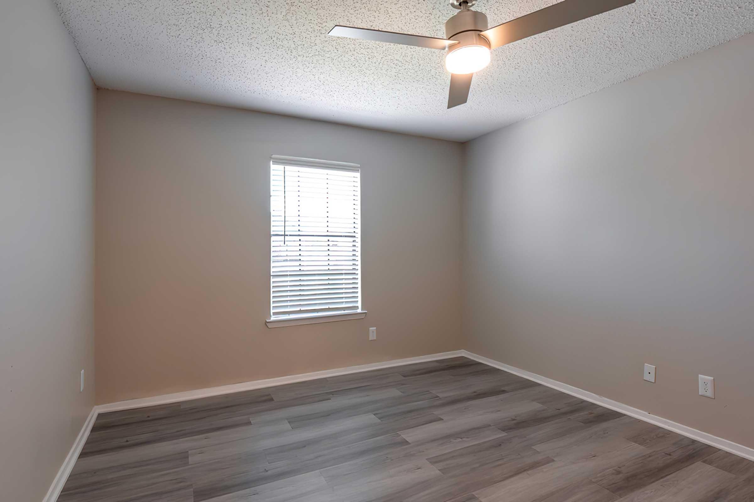 A well-lit, empty room with gray walls and a ceiling fan. There is a window with blinds allowing natural light to enter. The floor features light wood or laminate flooring, and the room has a clean, spacious feel with no furniture present.