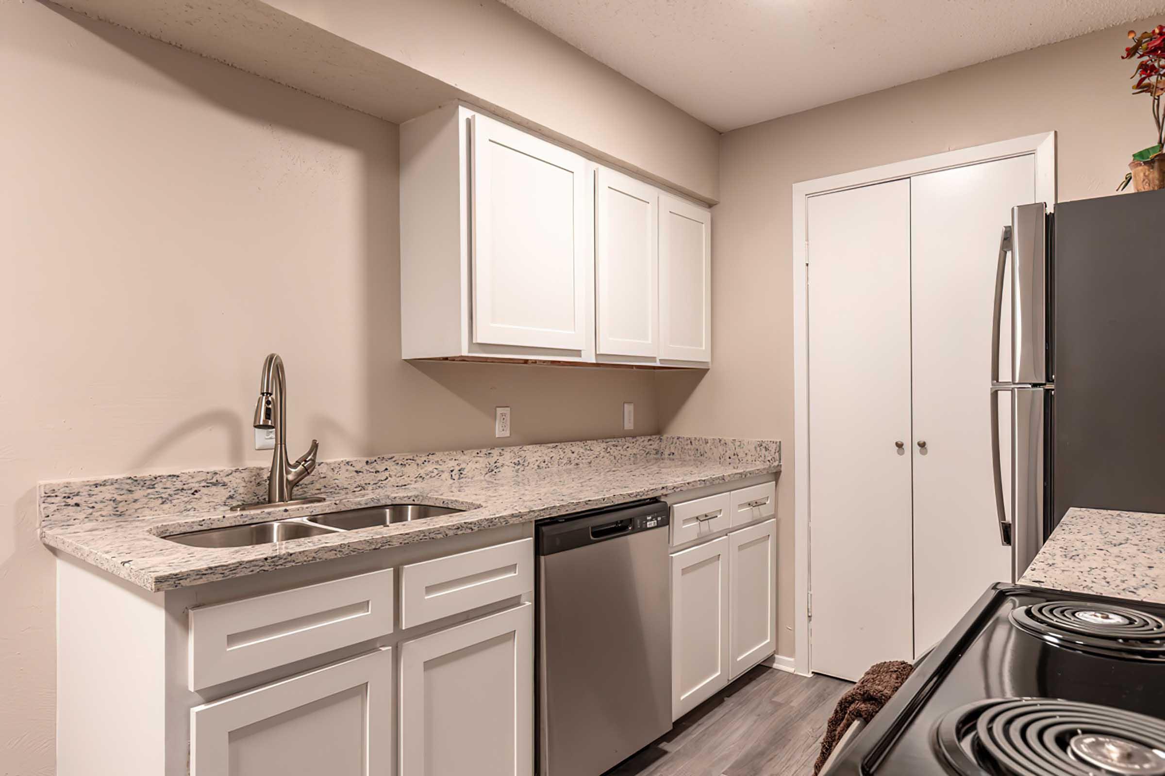 Modern kitchen featuring white cabinetry, a granite countertop, a double sink with a sleek faucet, a stainless steel dishwasher, and a black refrigerator. The space is well-lit and has an organized layout with a smooth electric cooktop and oven. Neutral wall colors enhance the contemporary aesthetic.