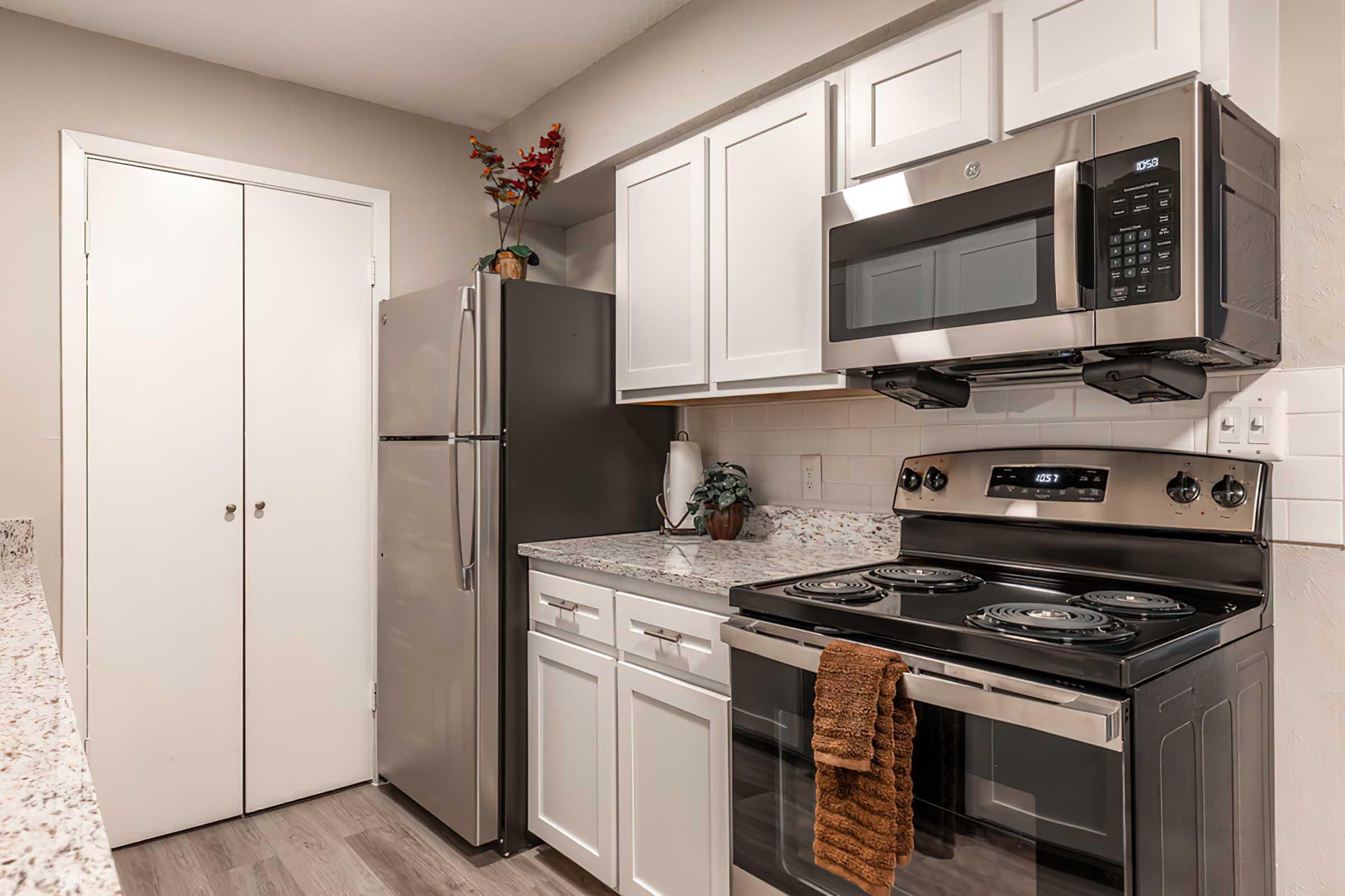 A modern kitchen featuring white cabinets, a stainless steel refrigerator, and a microwave above a black stove. The countertop is made of granite with a textured pattern. A decorative plant and a towel hang from the stove, and there is a closed sliding door visible in the background.