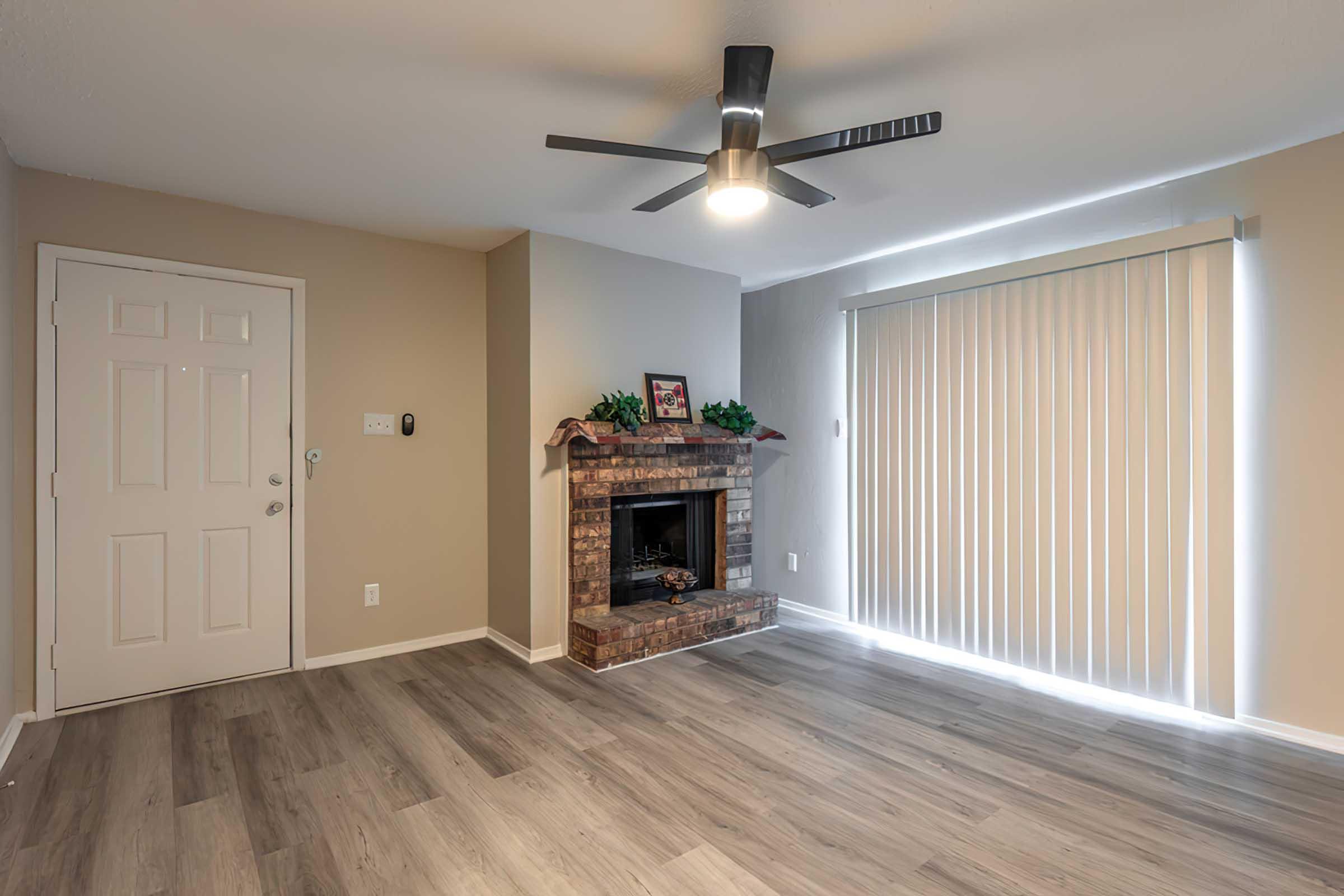 A cozy living room featuring a brick fireplace, a ceiling fan with light, and large sliding glass doors with vertical blinds. The walls are painted in neutral tones, and the flooring is light wood. A front door is visible on the left, adding to the inviting atmosphere of the space.