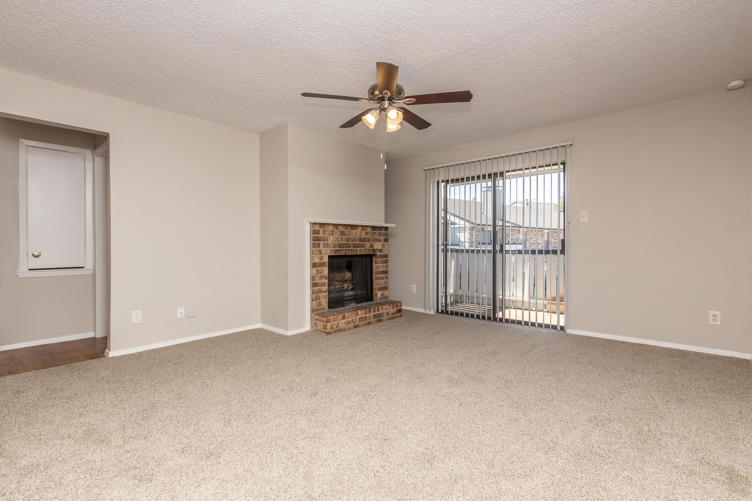 A spacious living room with light beige walls and carpet. It features a ceiling fan, a brick fireplace on one wall, and large sliding glass doors that open to a balcony. There is a small nook to the left with a door, adding character to the layout. The area is well-lit and inviting.
