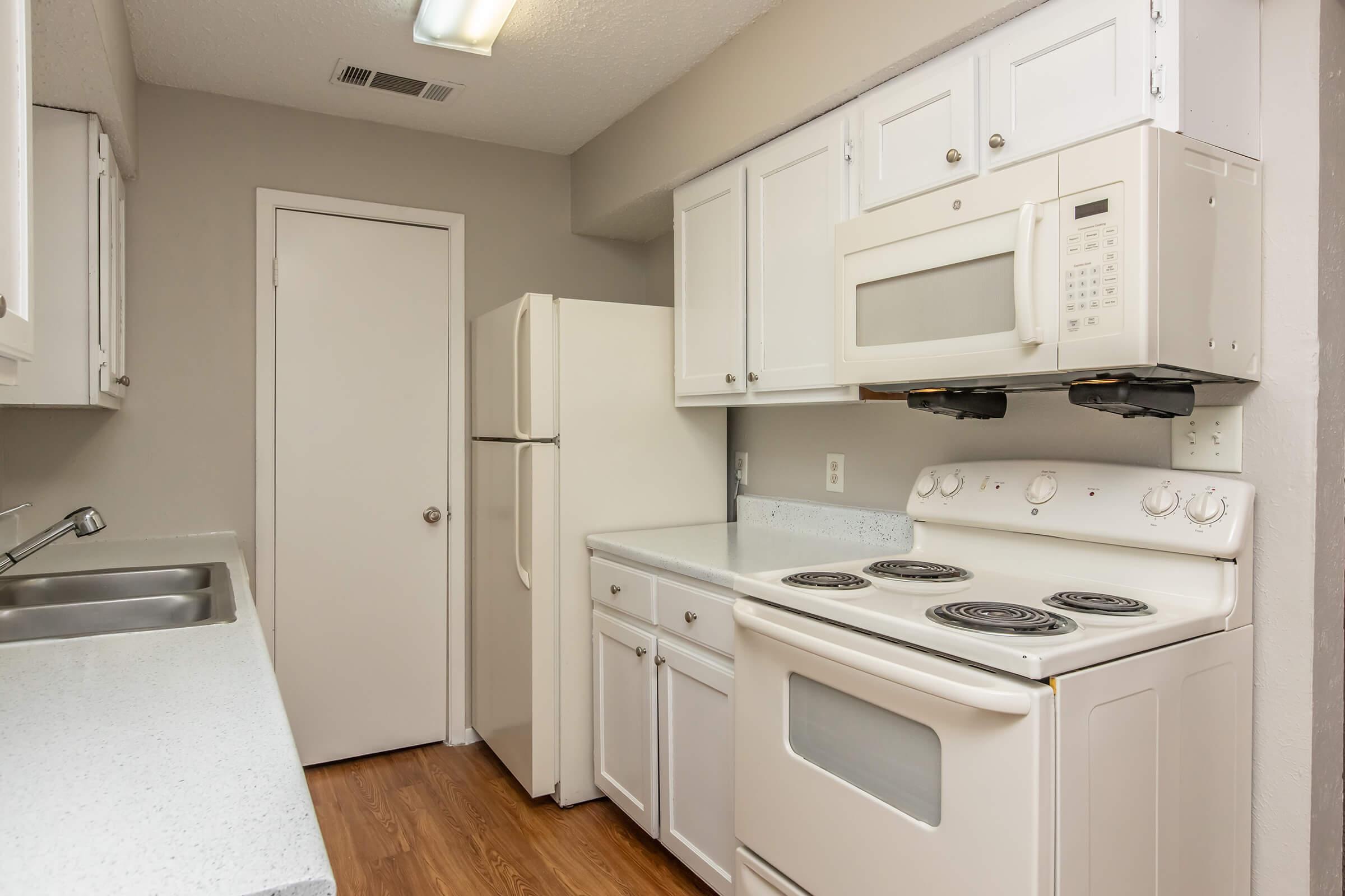 A small, modern kitchen featuring white cabinetry, a refrigerator, a microwave above the stove, and a four-burner electric range. The countertops are light-colored with a seamless design, and the flooring has a warm, wood-like appearance. The walls are painted in a neutral tone.