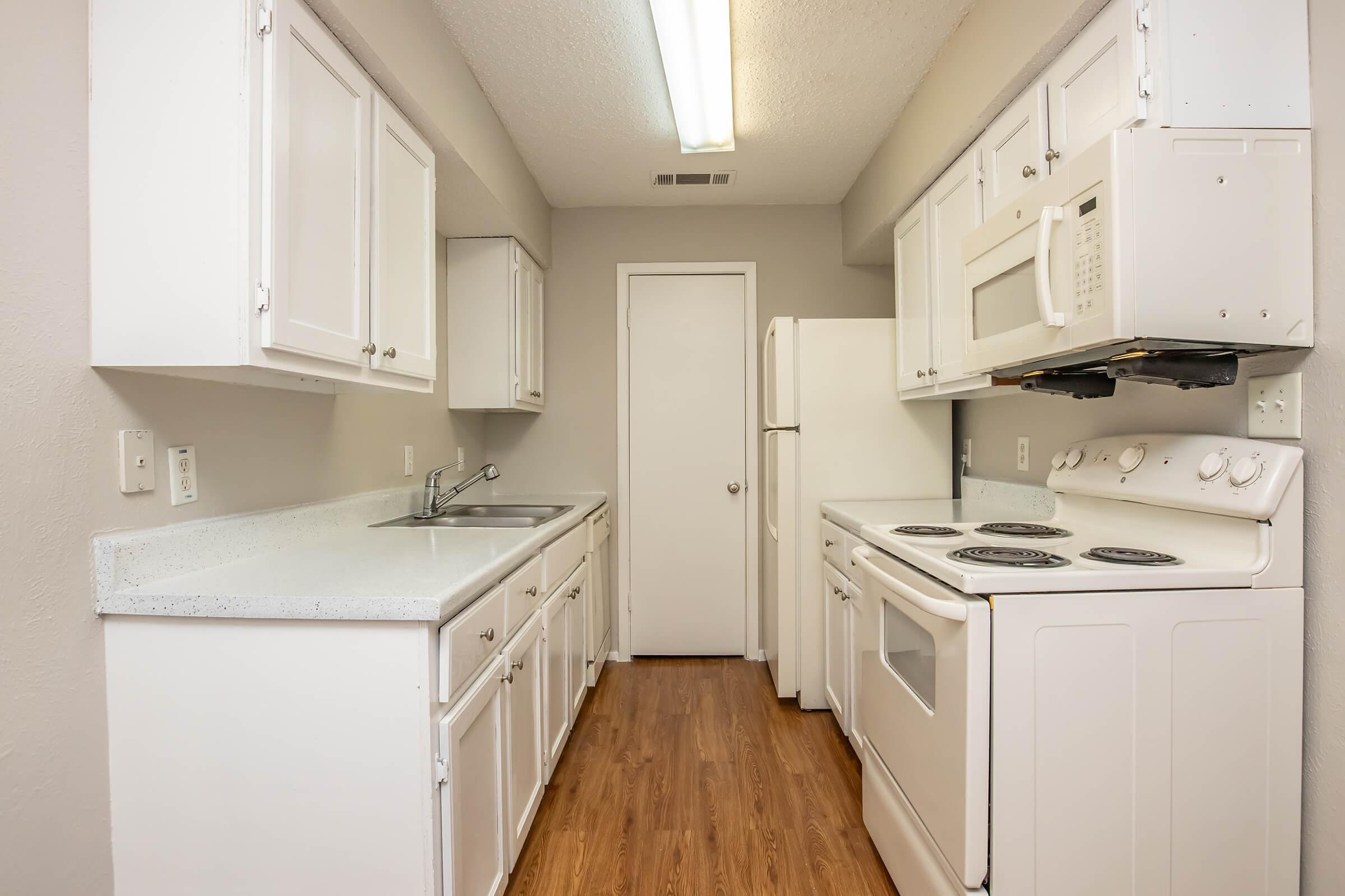 A clean and modern kitchen featuring white cabinetry, a stainless steel sink, an electric stove, and a microwave. The room has light-colored walls and wooden flooring, with an adjacent door leading to another area. Natural light is provided by a fluorescent ceiling fixture.