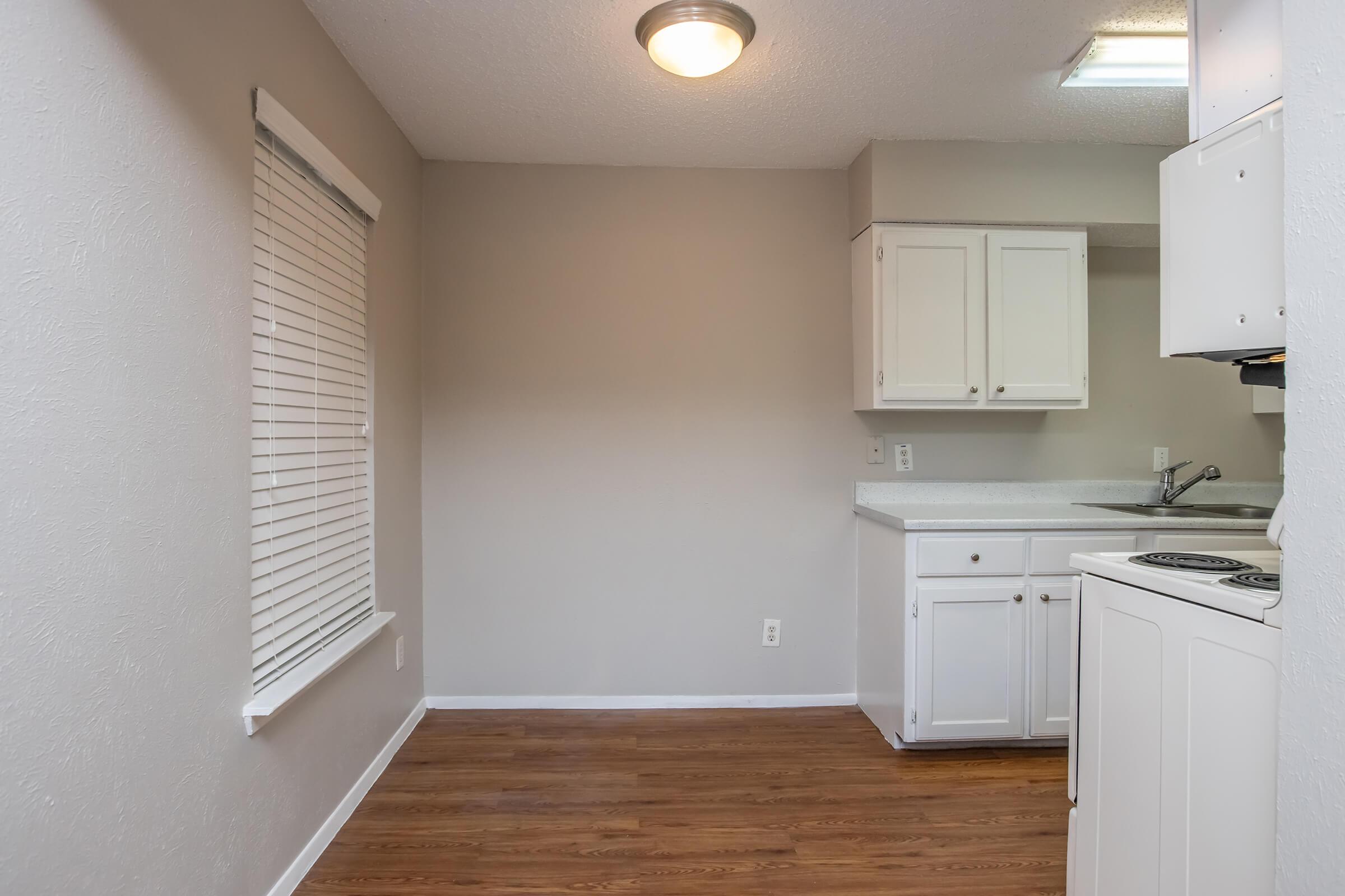 A small, empty kitchen area featuring white cabinets, a white countertop, and a sink. The walls are painted in a neutral color, and there is a window with blinds allowing natural light. The floor is made of wooden planks, and the space appears clean and bright, suitable for a compact living environment.