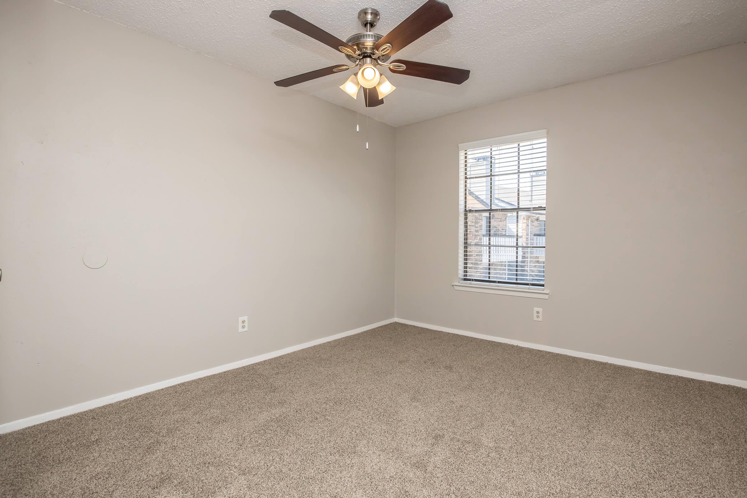 Empty room with light beige carpet, a ceiling fan with four blades, and a window with blinds letting in natural light. The walls are a light gray color, and there is a baseboard along the floor. The space is uncluttered and ready for furnishing.