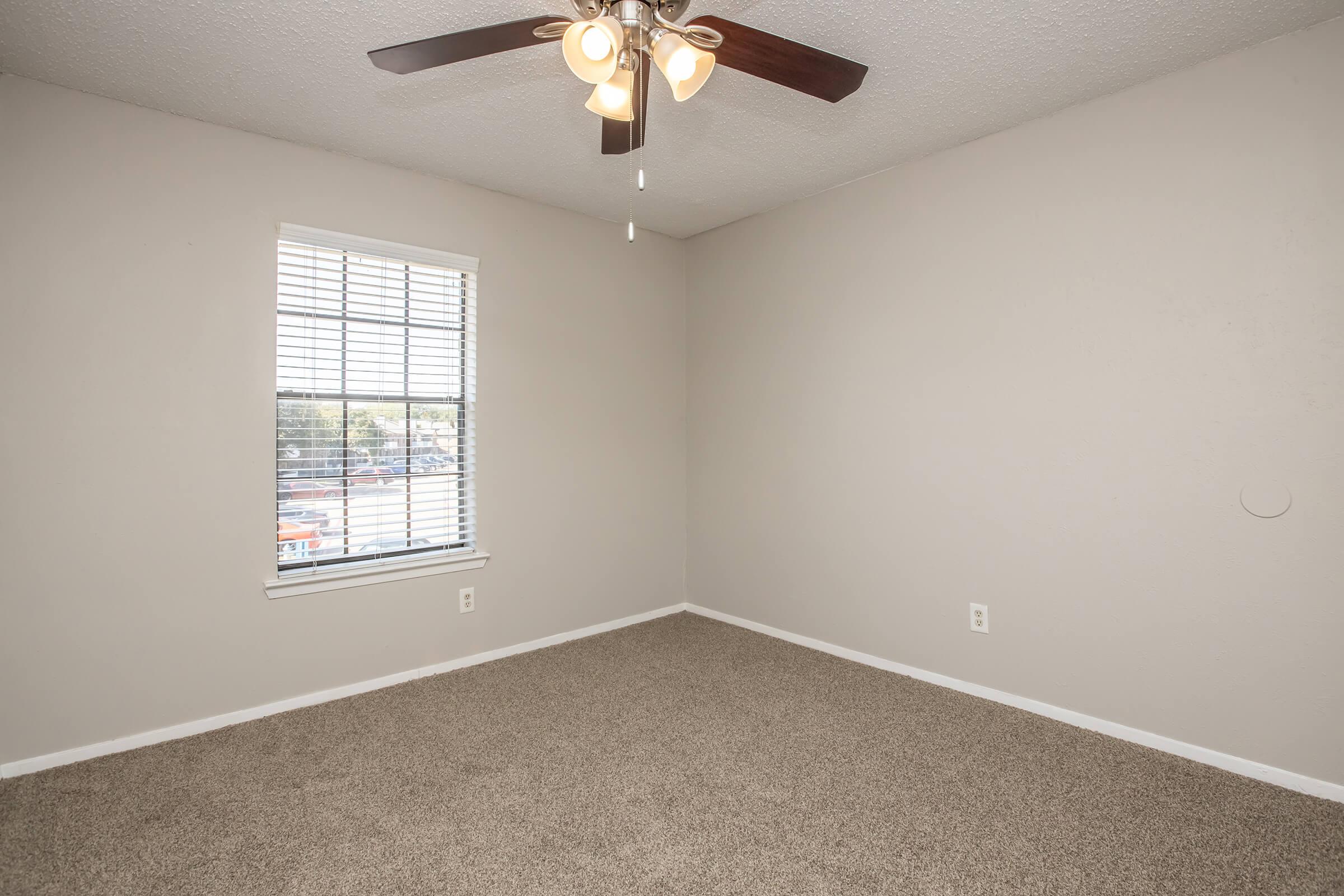 Empty room with light beige walls and a light brown carpet. A ceiling fan with three lights hangs from the ceiling. A window with white blinds allows natural light to enter, revealing an outdoor view. The space is uncluttered and awaiting furnishings.